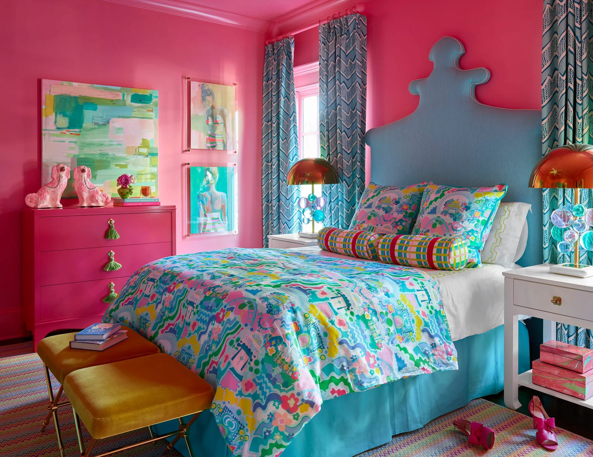A maximalist bedroom features fuchsia walls, a tall light blue upholstered headboard, colorful patterned bedding, and a matching pink dresser.