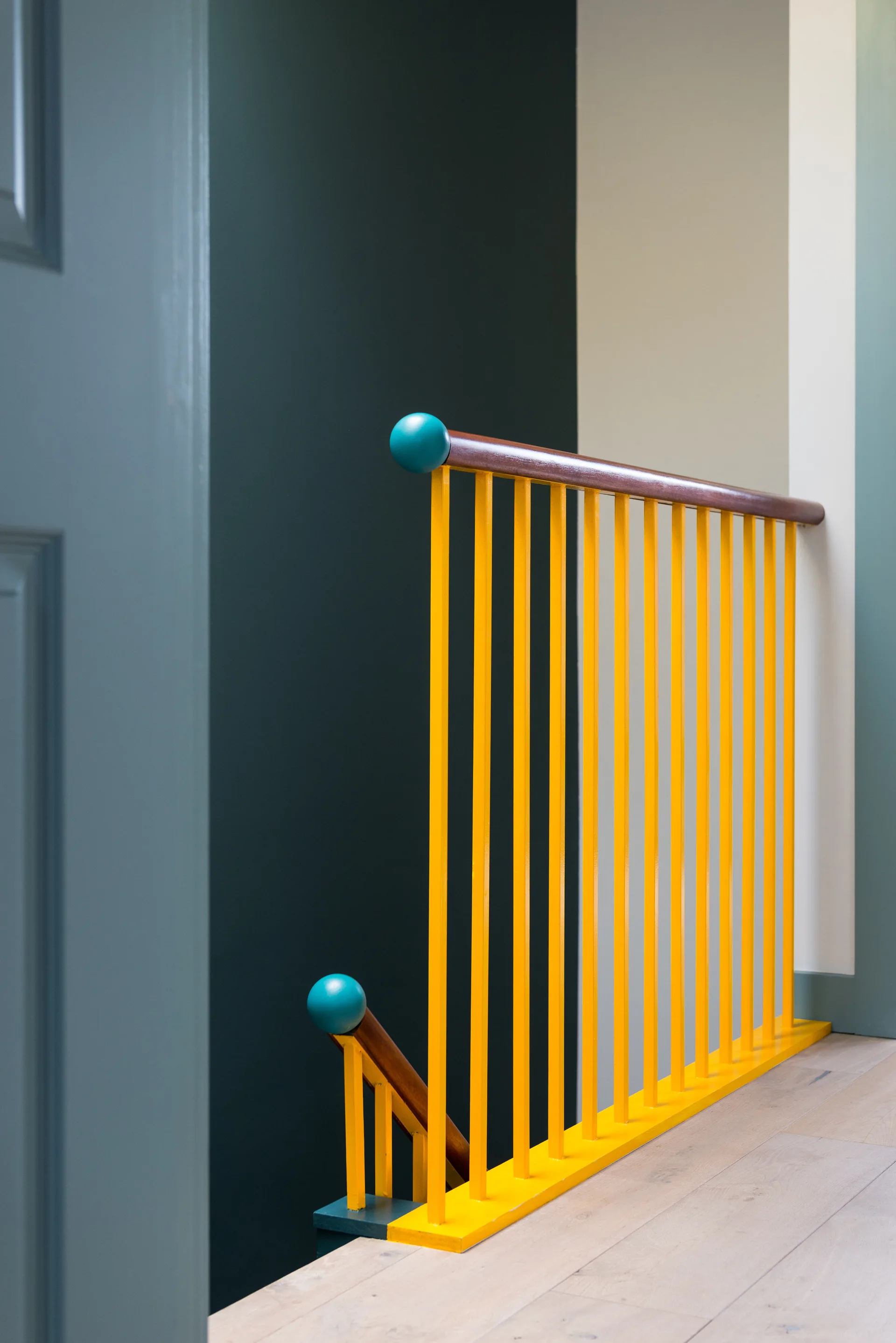 A staircase landing showing bright yellow spindles, a wooden handrail with teal spherical finials, and a dark green wall.