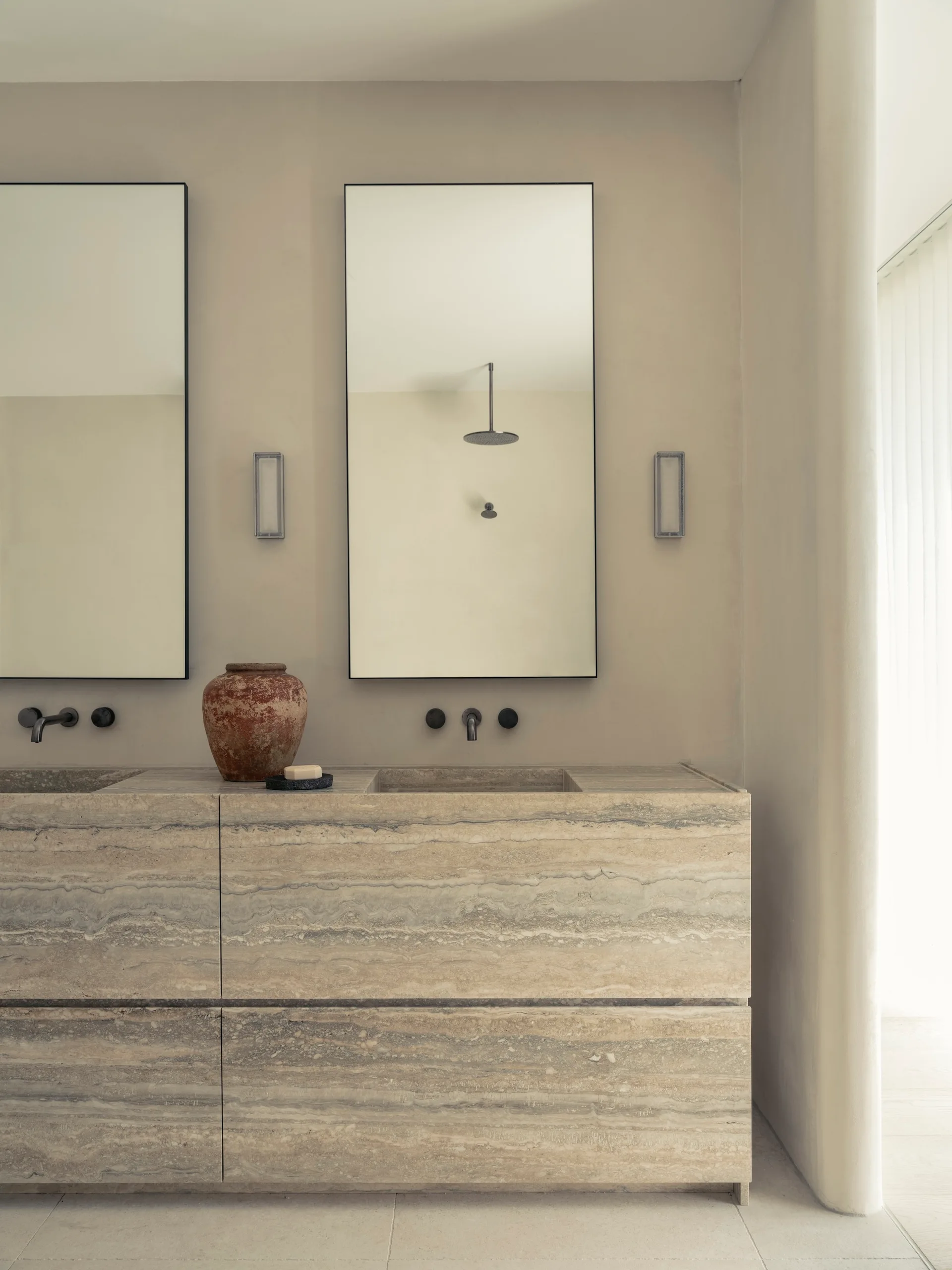 A beige plaster bathroom features a travertine stone double vanity with integrated sinks, black wall-mounted faucets, and two rectangular mirrors.