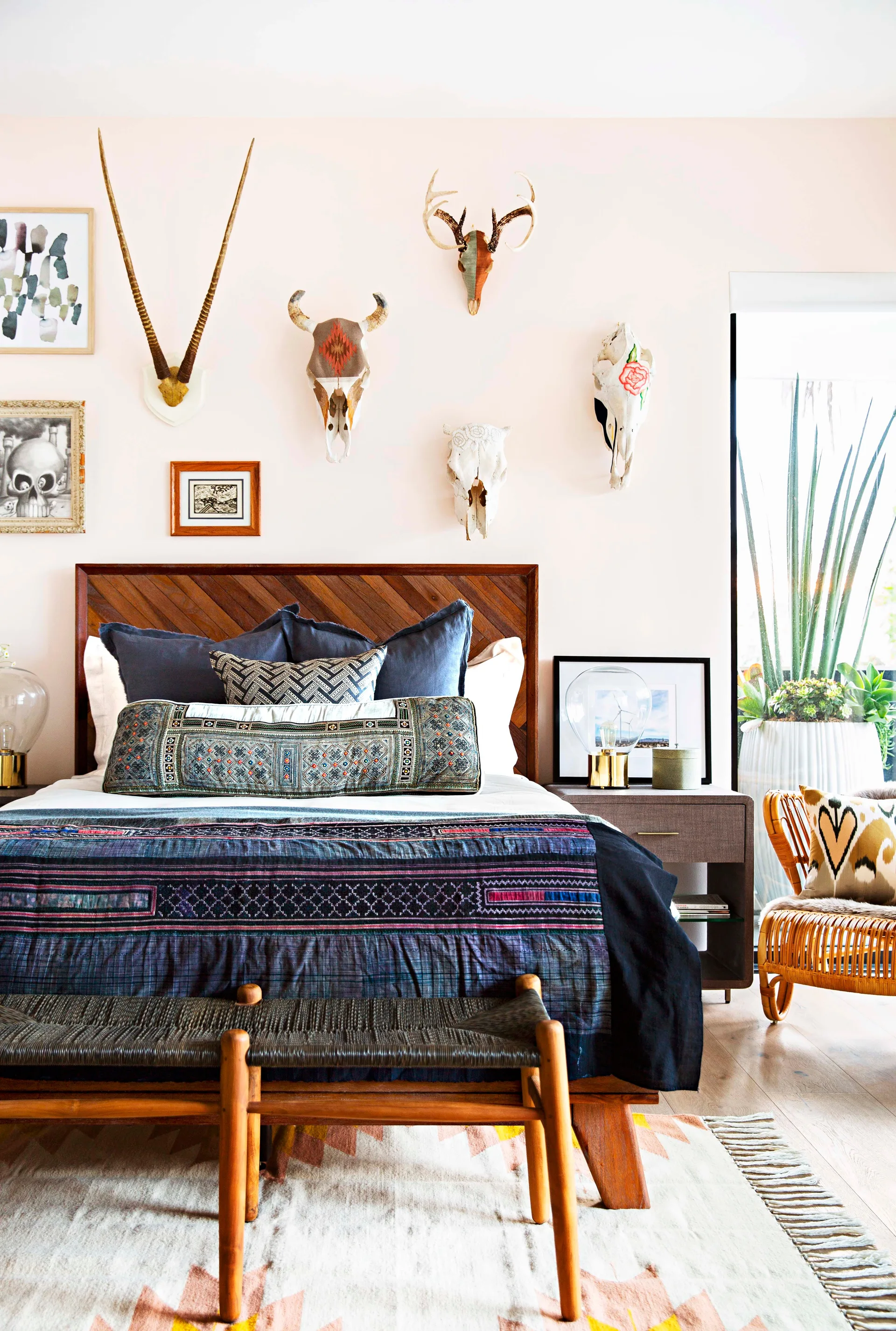 A bedroom with a wooden herringbone headboard, pale pink walls decorated with animal skulls, patterned blue bedding, and a wood bench.