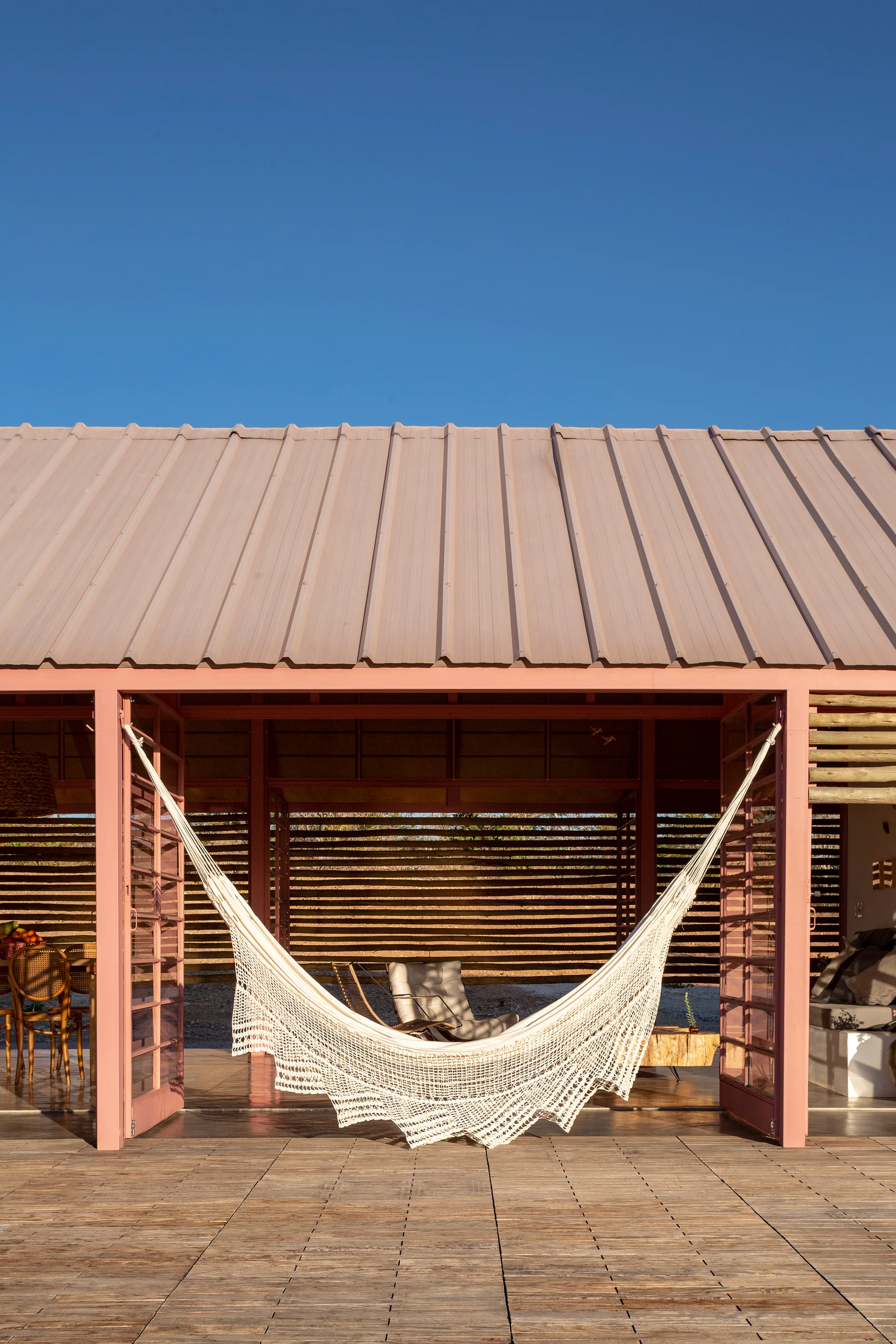 A white crocheted hammock hangs in the center of a dusty pink painted patio structure with wooden decking under a clear blue sky.