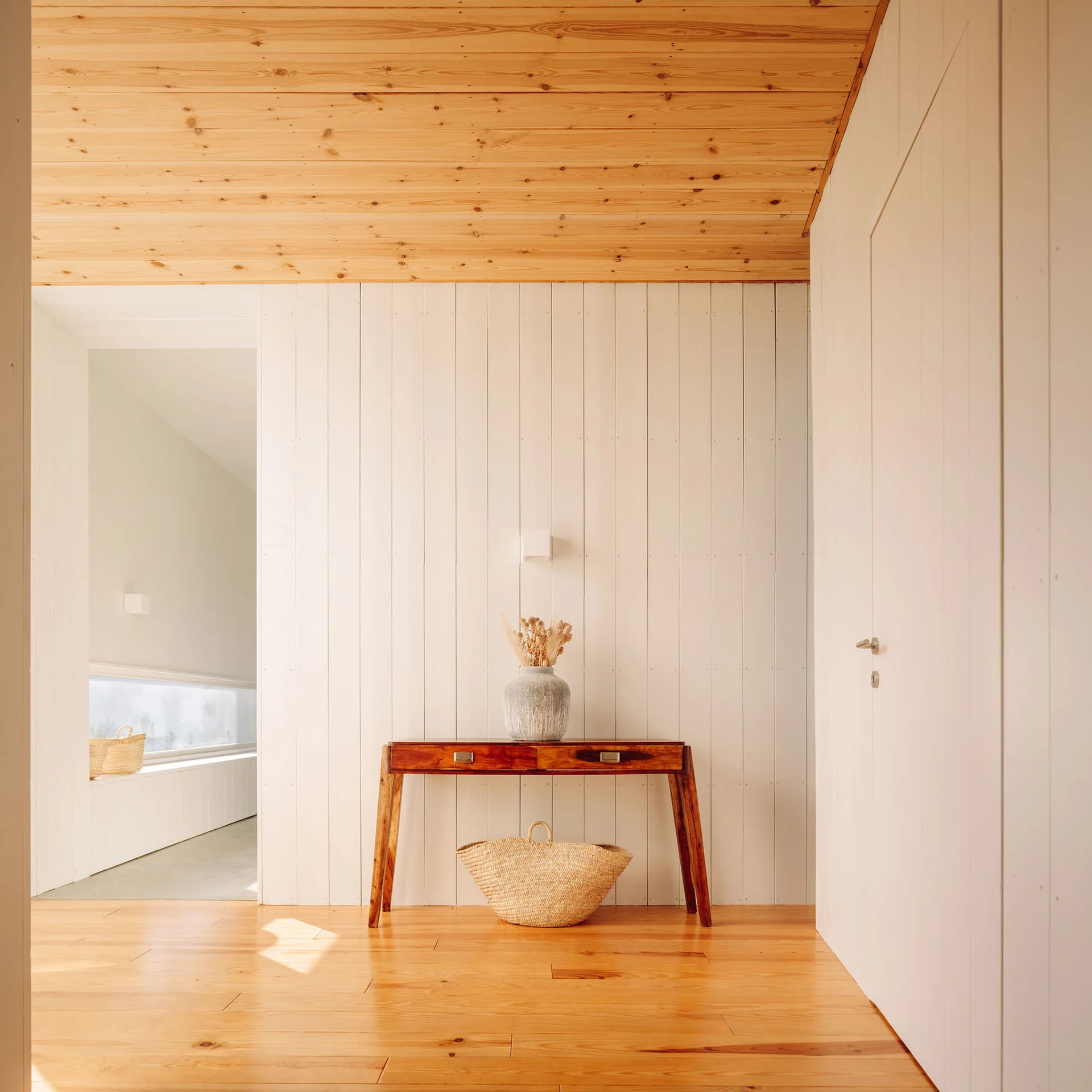 A minimalist hallway featuring a wooden console table against white vertical plank walls, framed by a natural wood floor and ceiling.