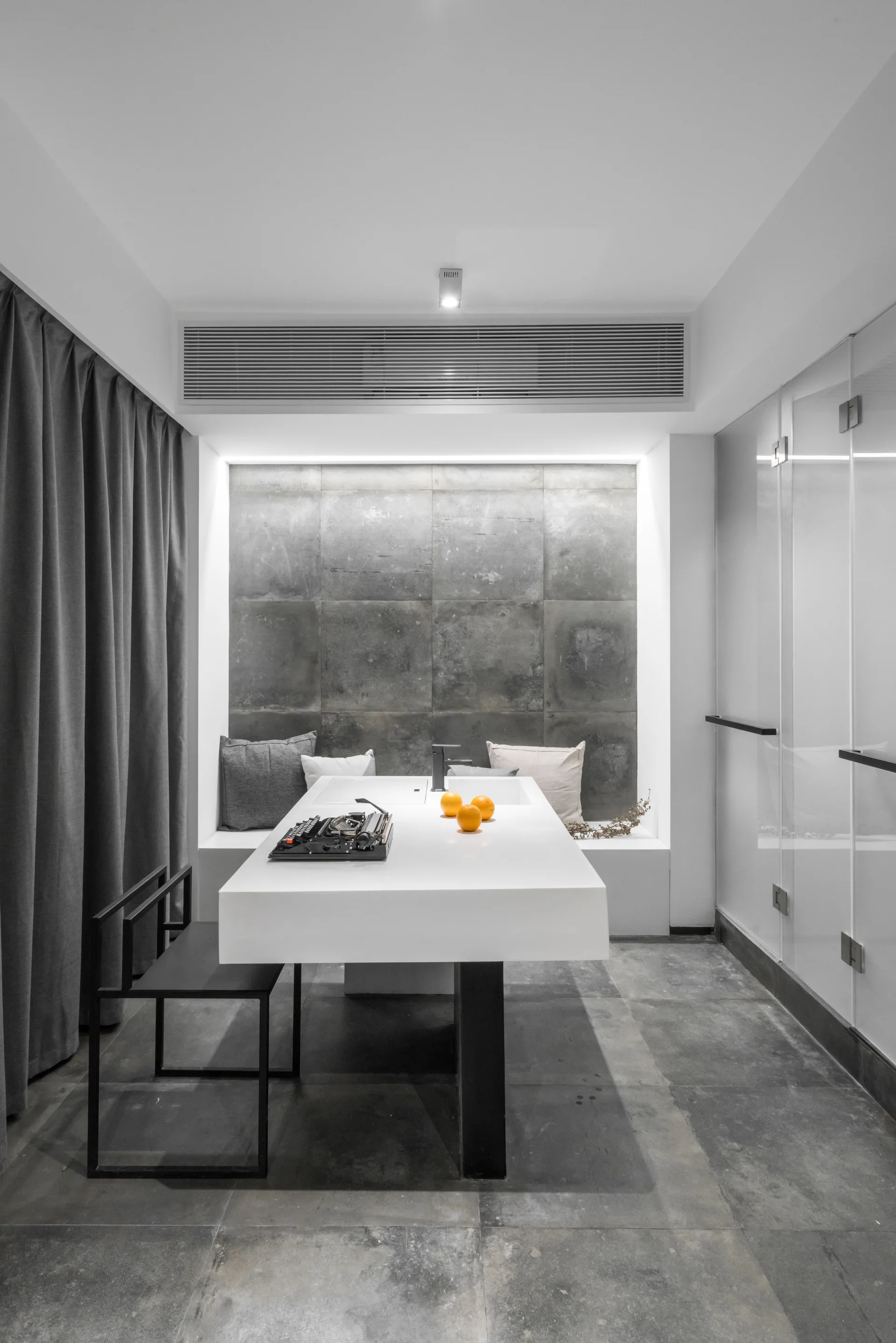 A minimalist white kitchen island with a integrated faucet sits before a grey tiled wall with a built-in bench.