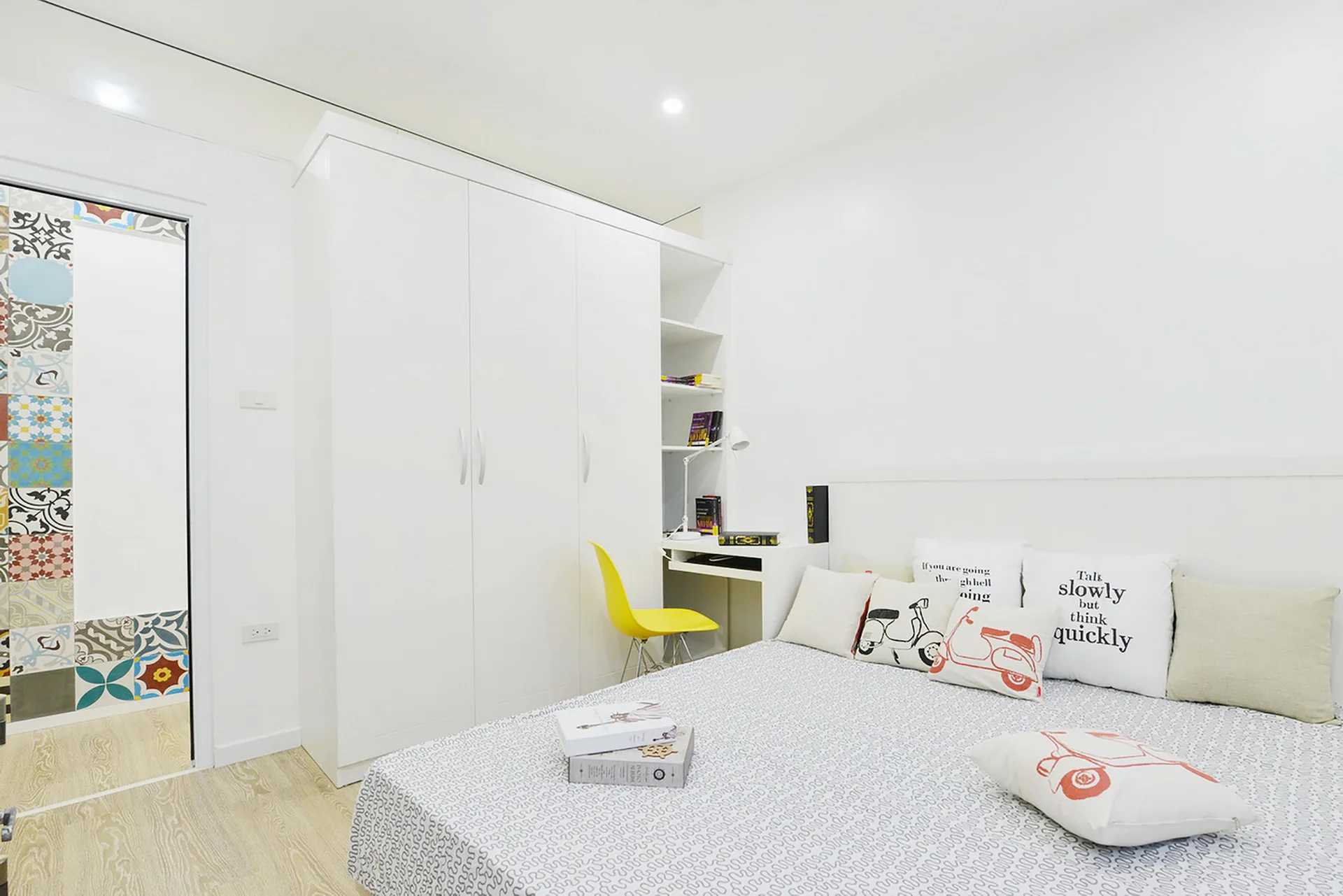 A bright white bedroom features a built-in wardrobe and desk unit with a yellow chair, next to a bed decorated with printed pillows.