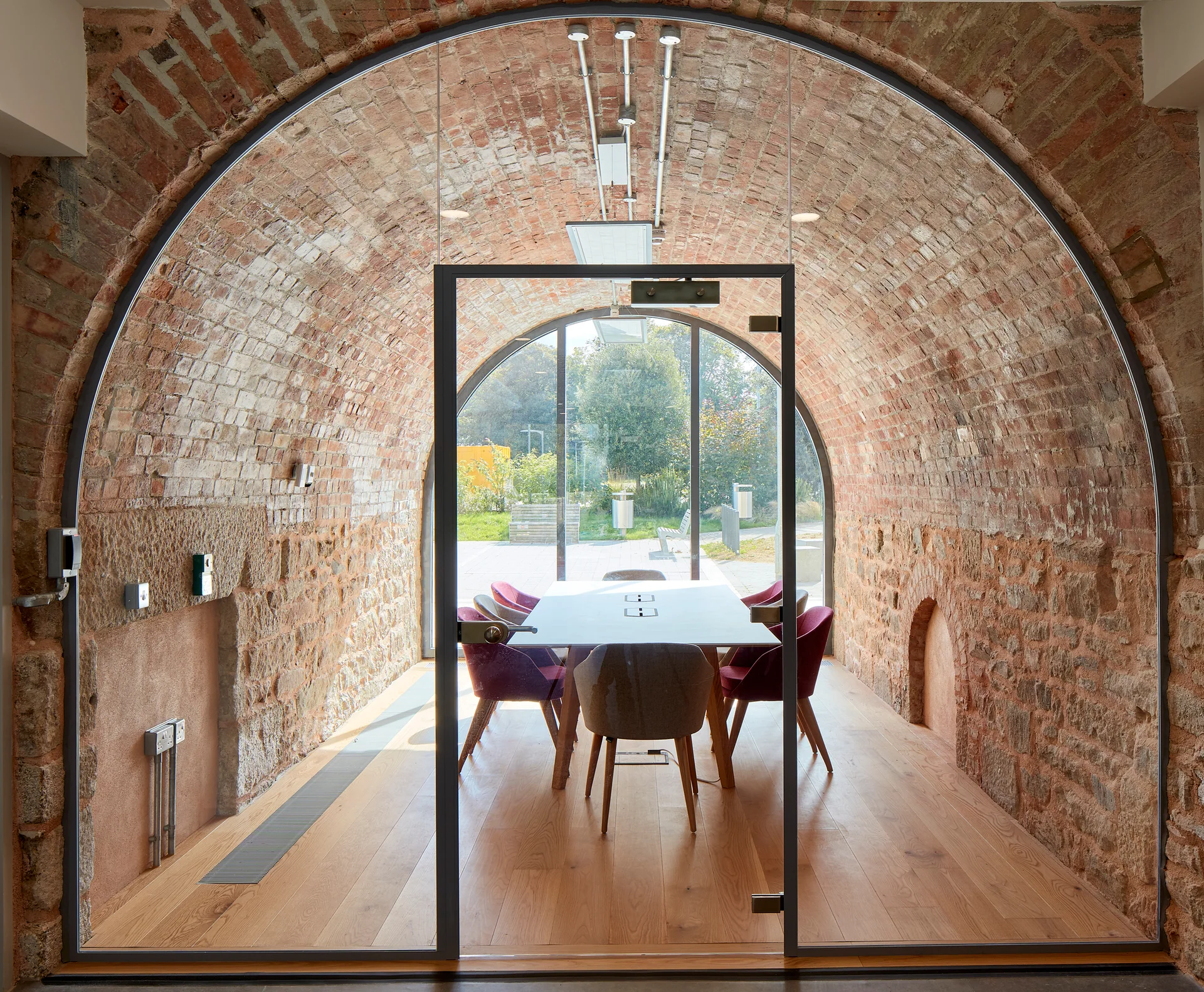 A conference table with colorful chairs sits inside a historic arched brick vault behind a modern glass wall partition.