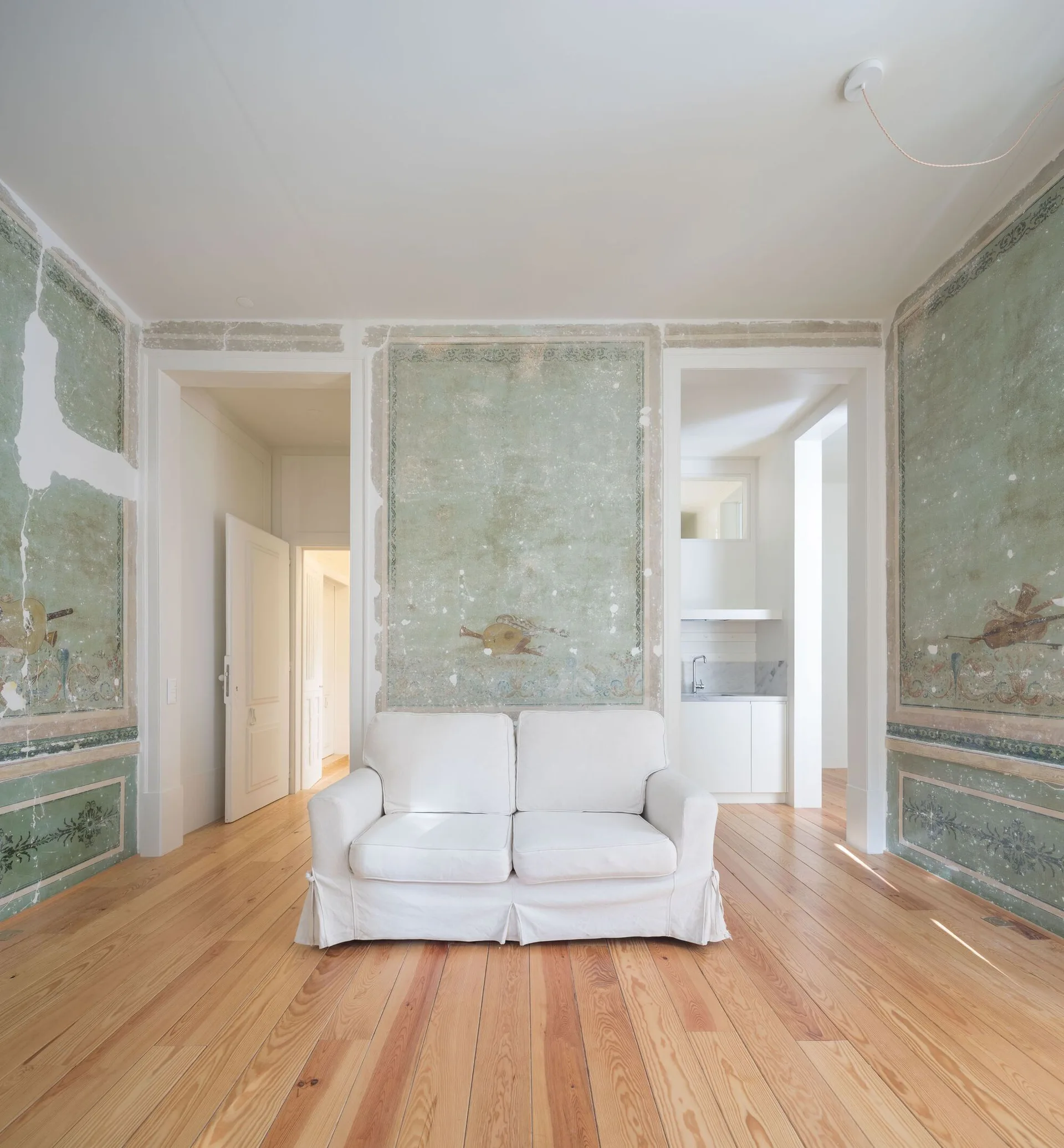 A white slipcovered sofa sits on natural wood floors in a room with distressed sage green fresco mural walls and open doorways.