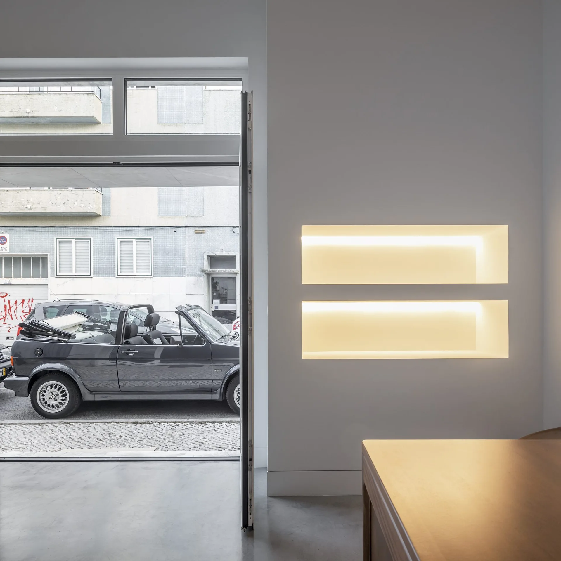 A minimalist white room with concrete floors features two stacked horizontal wall niches glowing with warm recessed lighting next to an open glass door.