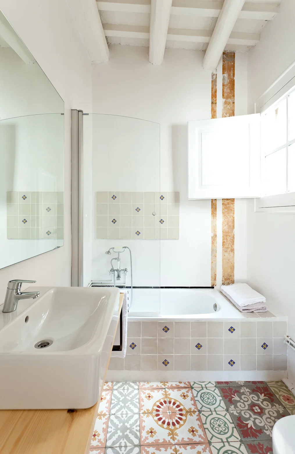 A bright rustic bathroom with decorative patterned floor tiles, a white bathtub, and a wood vanity with a ceramic basin.