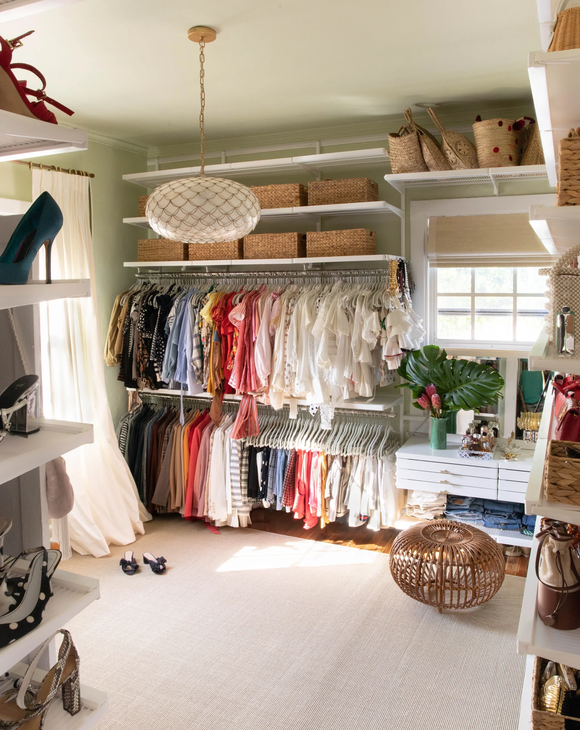An organized walk-in closet with sage green walls, white open wire shelving, hanging clothes, woven storage baskets, and a round wicker ottoman.