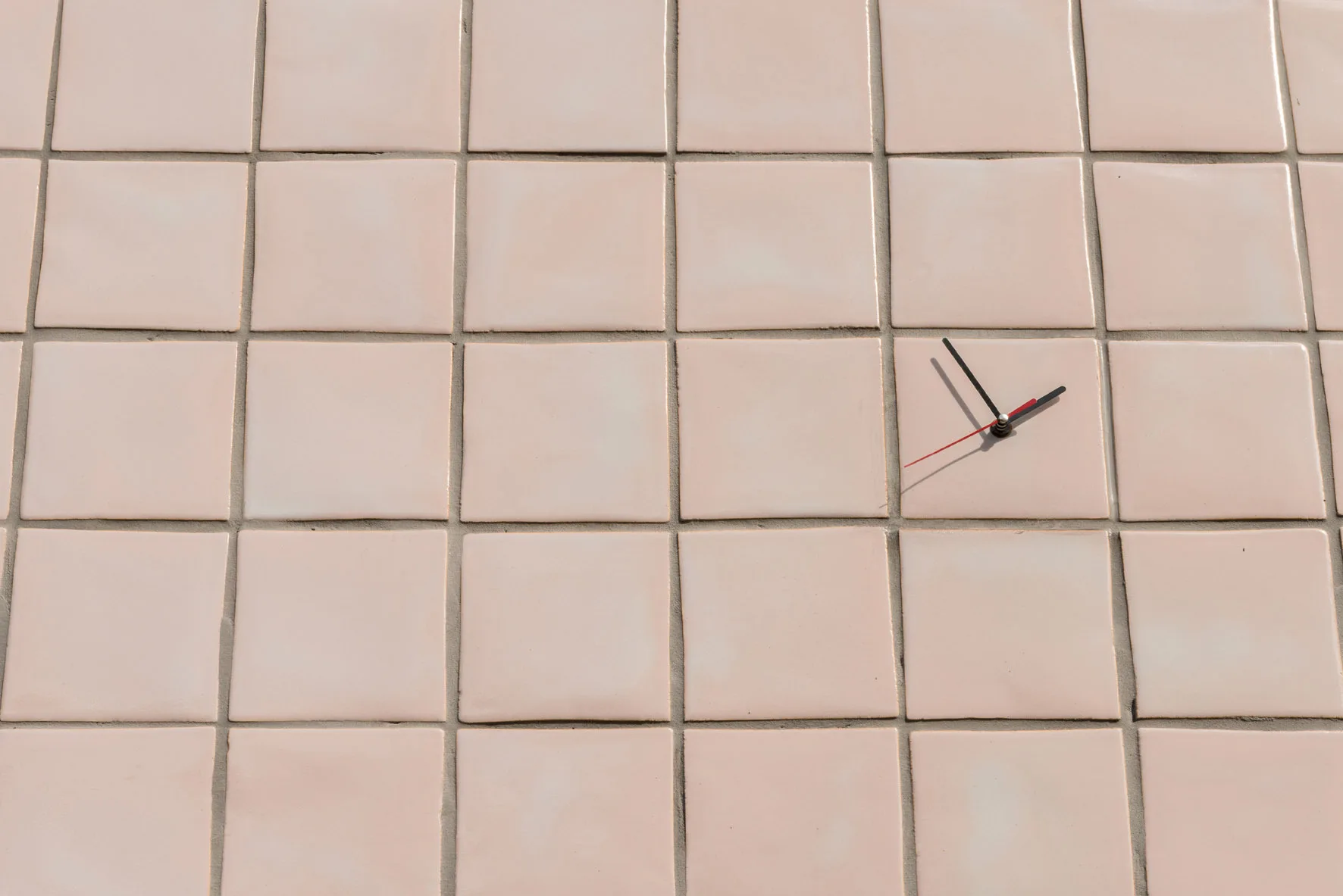 A close-up view of light pink square ceramic tiles on a wall with simple black and red clock hands mounted directly onto one tile.