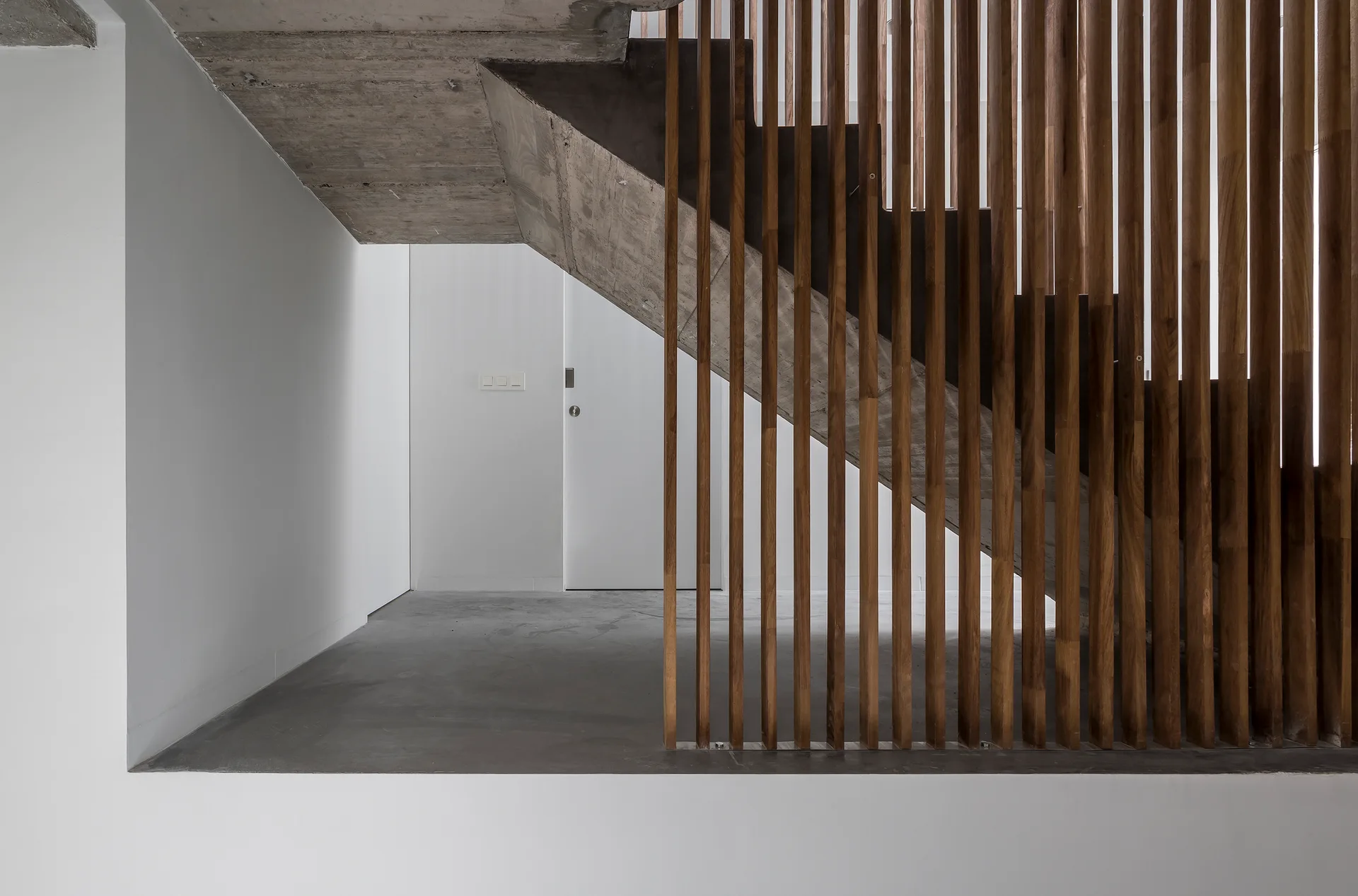 An interior hallway features an exposed concrete staircase enclosed by a vertical wooden slat balustrade next to a white wall.