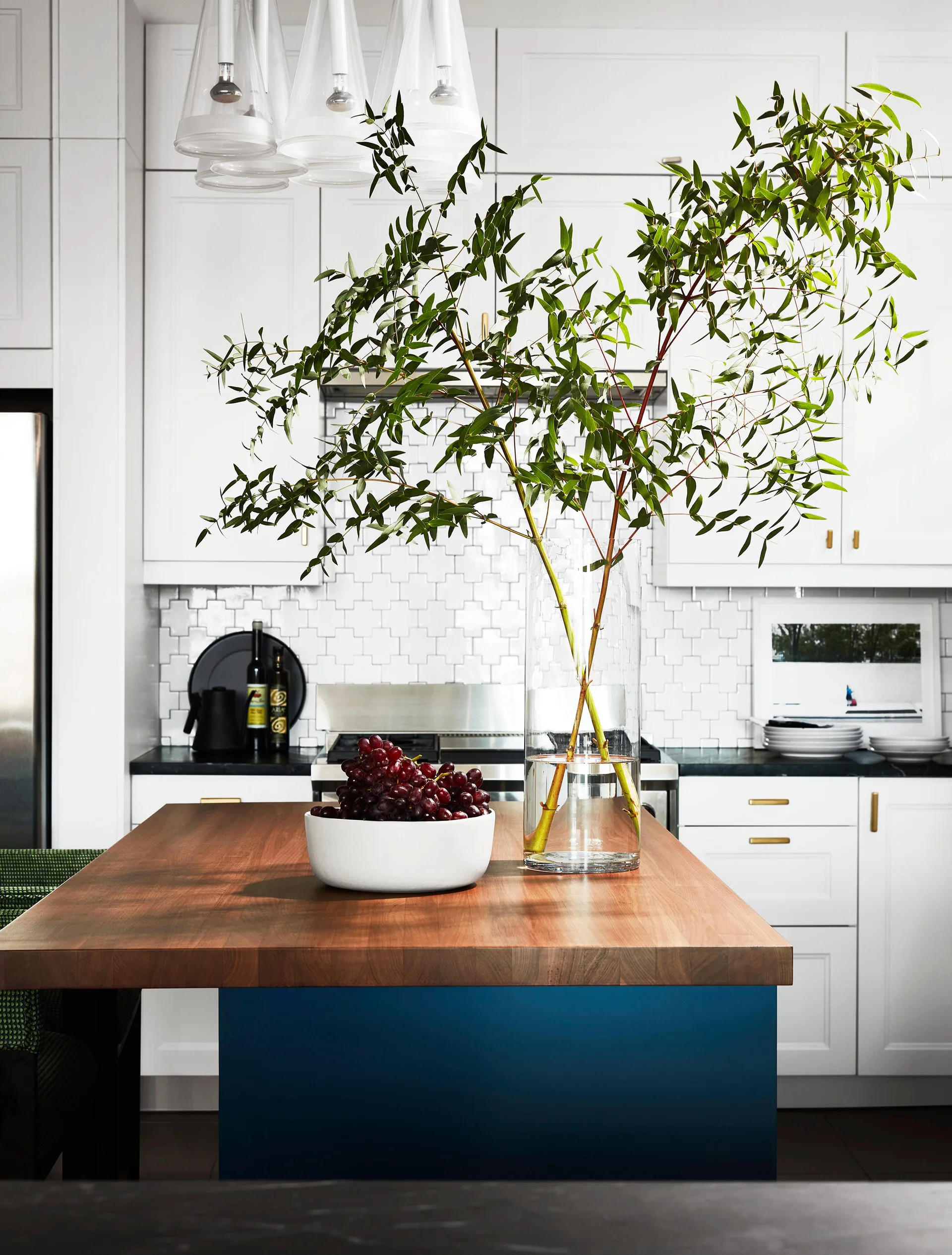 A kitchen island with a wood countertop and blue base holds a bowl of grapes and a glass vase with tall green branches in front of white cabinets.