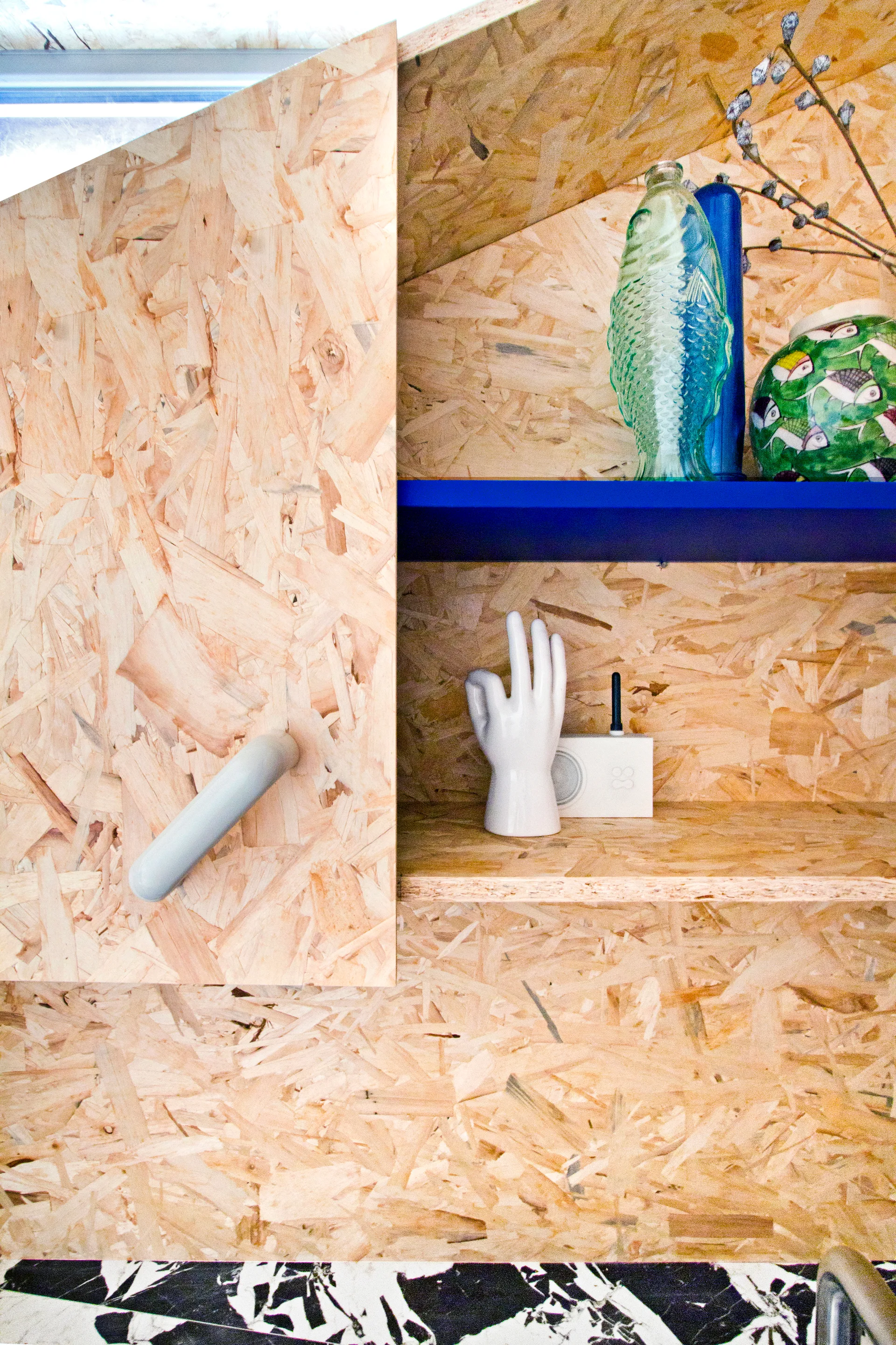 A close-up of a custom OSB cabinet featuring an open storage nook with a bright blue shelf, ceramic hand sculpture, and fish-shaped glass bottle.