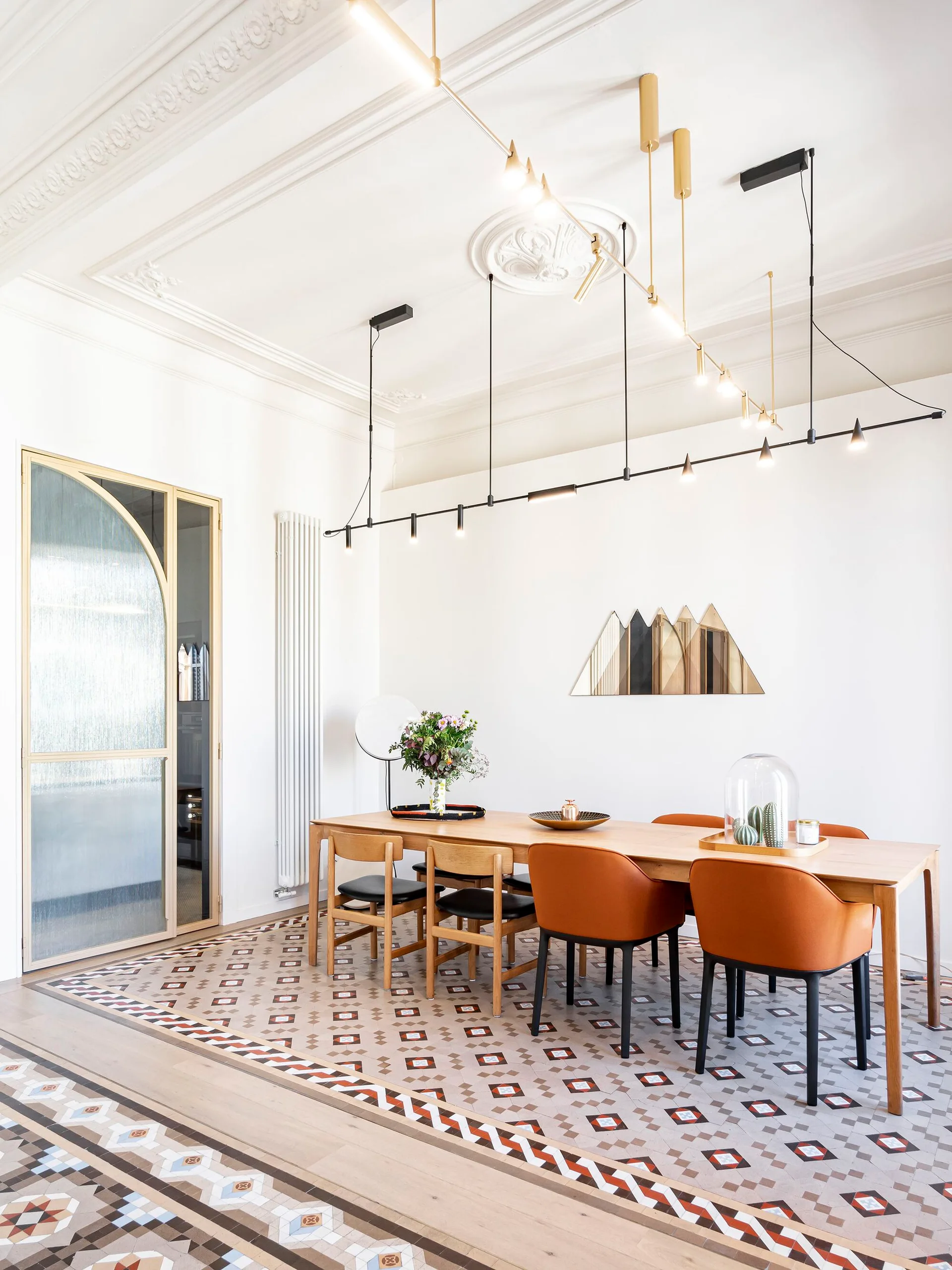 A bright dining room with a long wood table, orange and black chairs, patterned floor tile, and a black linear light fixture.