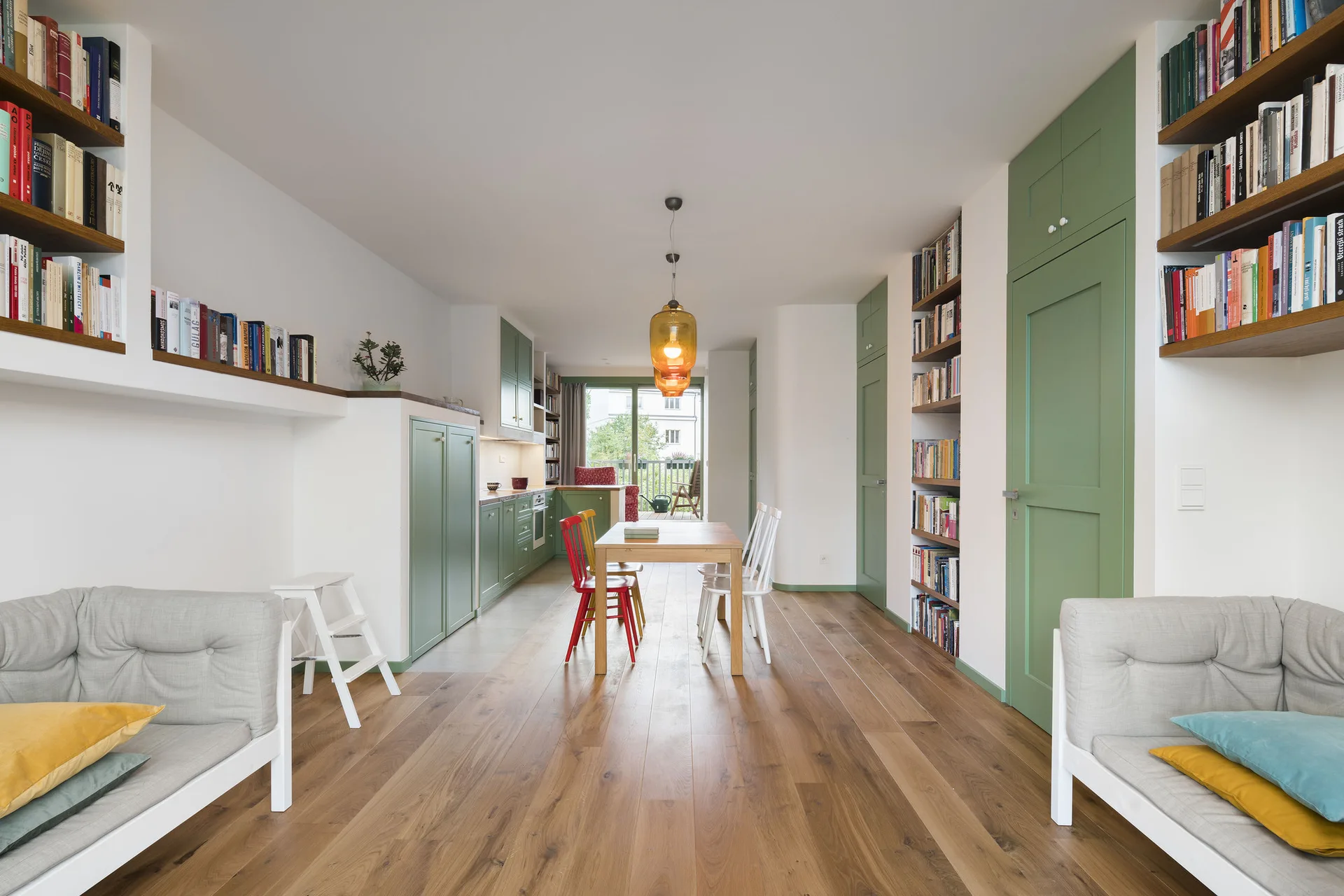An open-plan living and dining room features sage green cabinetry, built-in bookshelves, two grey sofas, and a central wooden dining table.