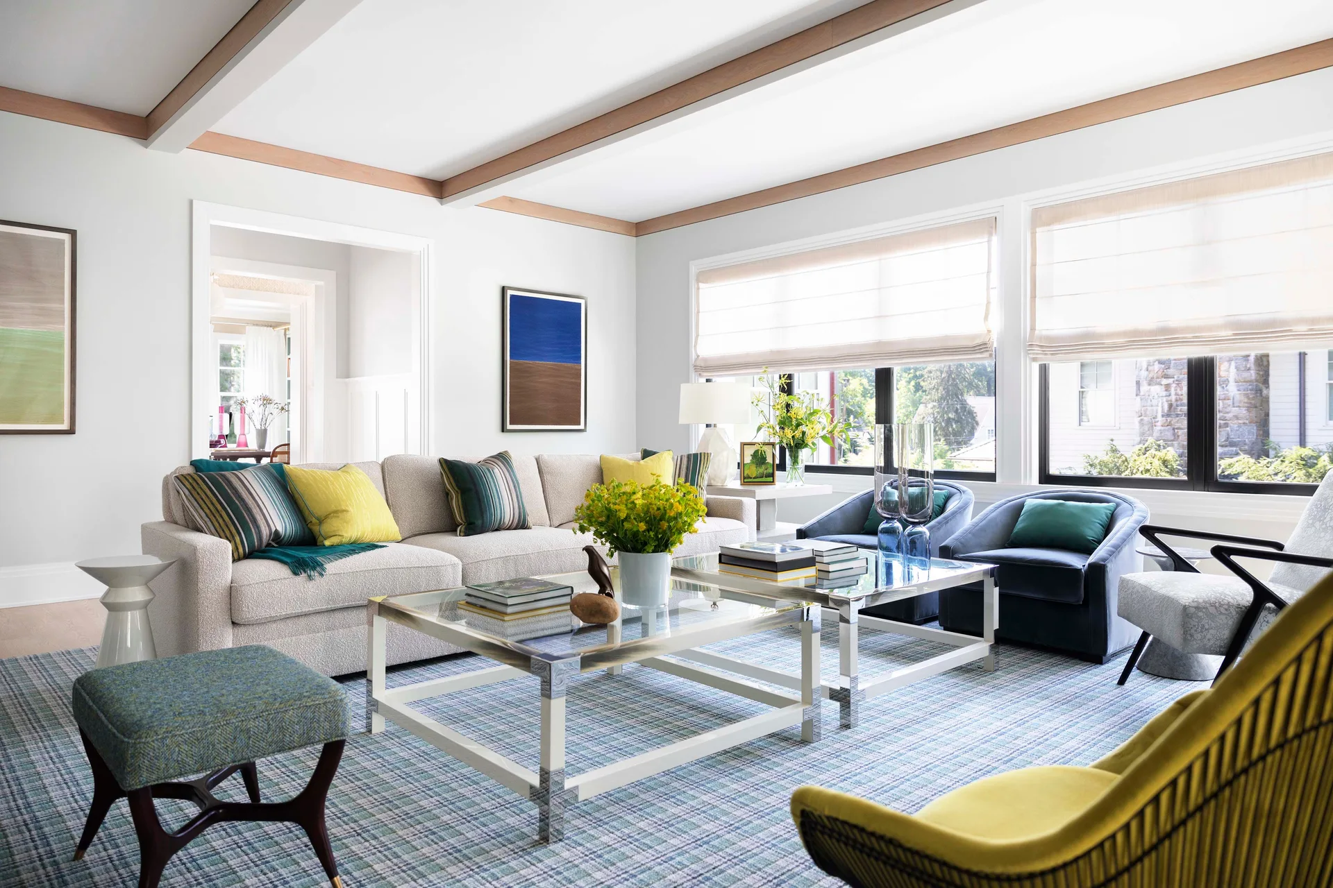 A bright transitional living room featuring a cream sofa, blue club chairs, and two glass coffee tables on a blue plaid rug.