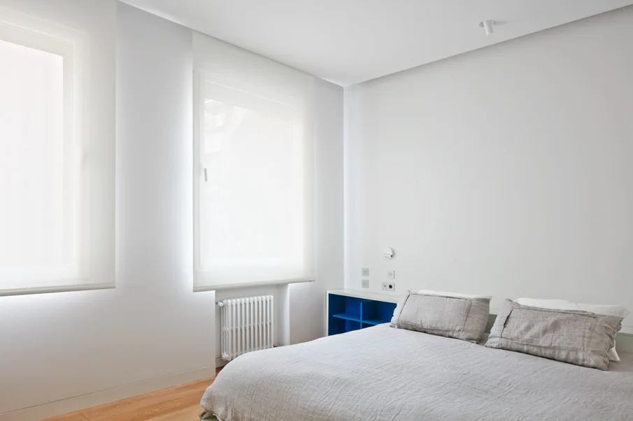 A minimalist white bedroom featuring a bed with neutral grey linen sheets, a vivid blue open bedside storage shelf, and windows with white roller shades.