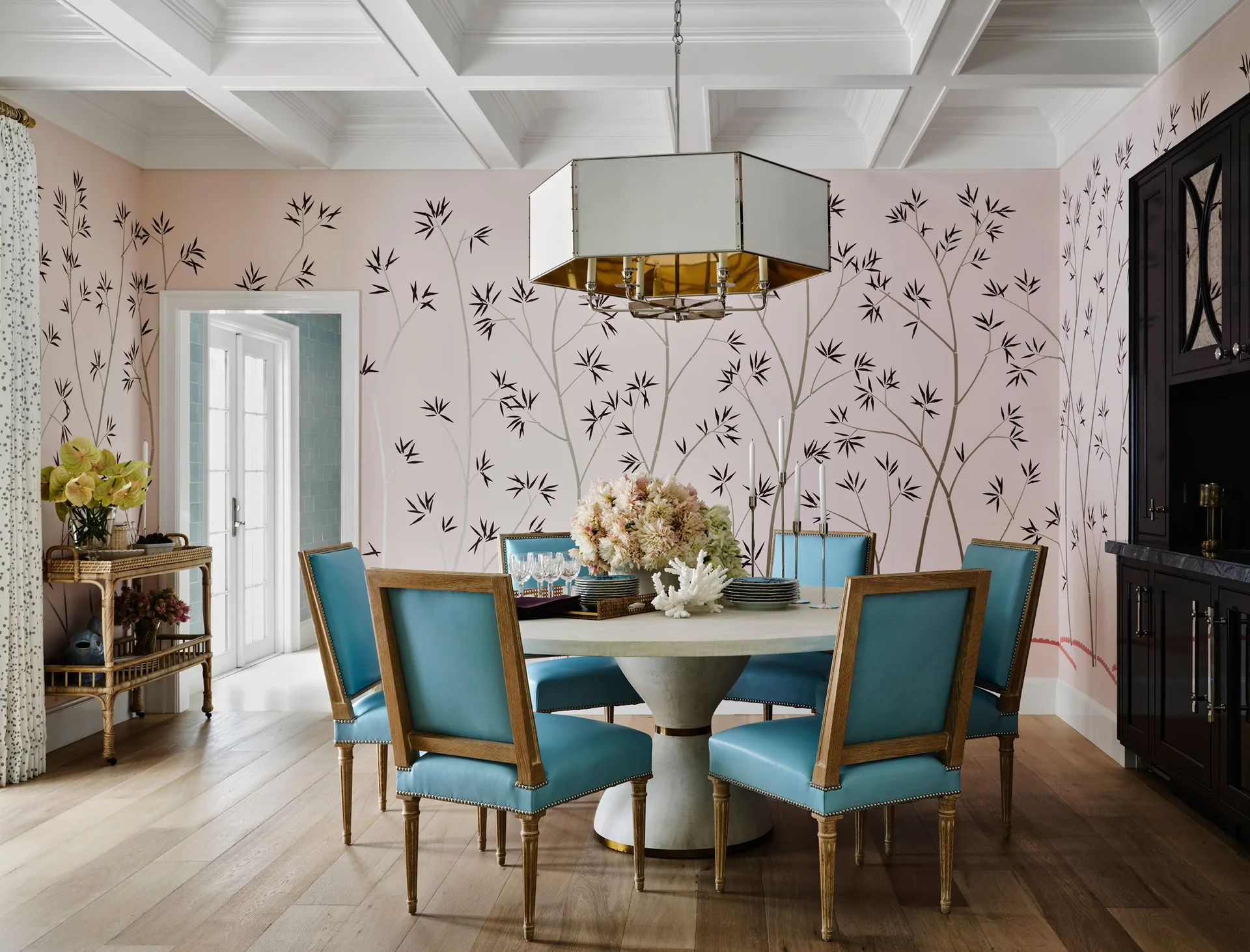 A dining room features a round pedestal table surrounded by bright blue chairs, pink wallpaper with a black bamboo pattern, and a coffered ceiling.