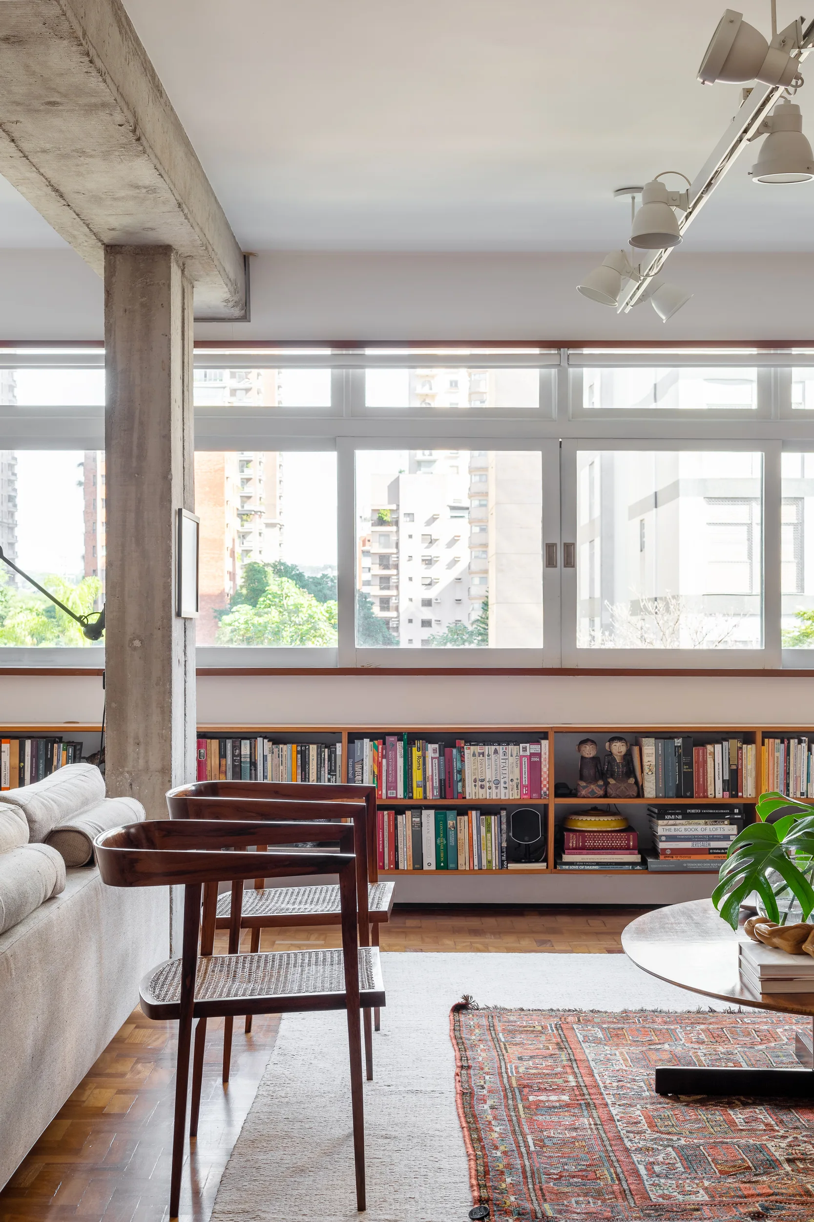 A modern living room featuring a long low wood bookshelf under large windows, an exposed concrete pillar, and wood and cane chairs.