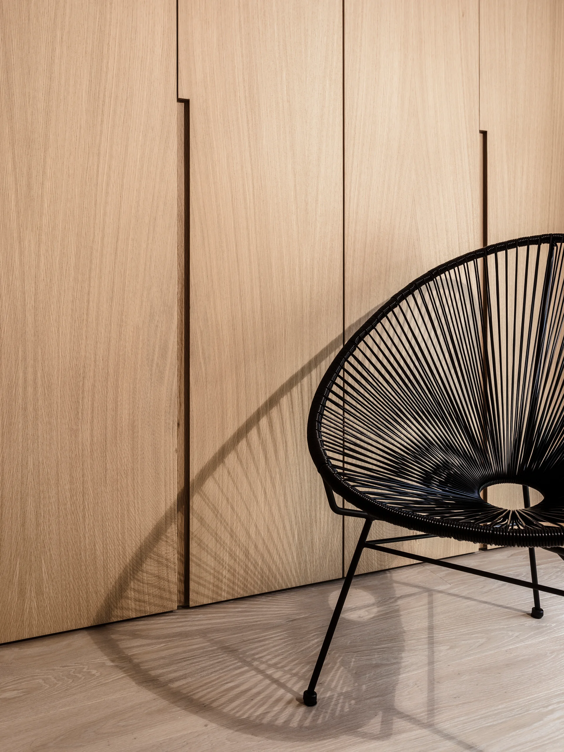 A black corded accent chair sits on a light oak floor against a wall of full-height vertical wood cabinetry.