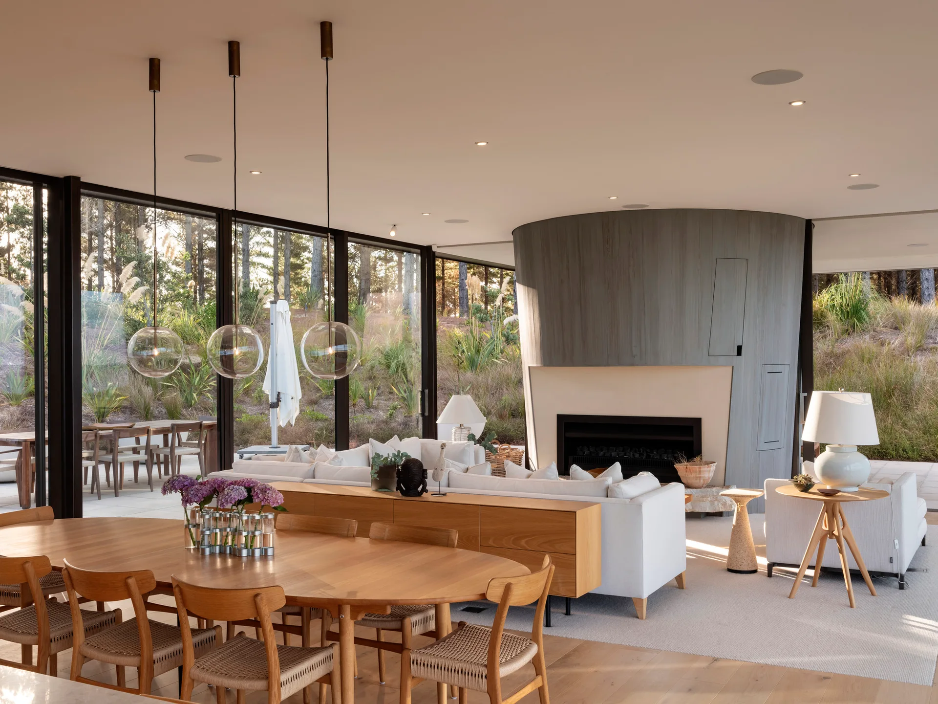 An open-plan interior features an oval wood dining table, a white sectional sofa, and a curved grey fireplace backdrop overlooking outdoor greenery.