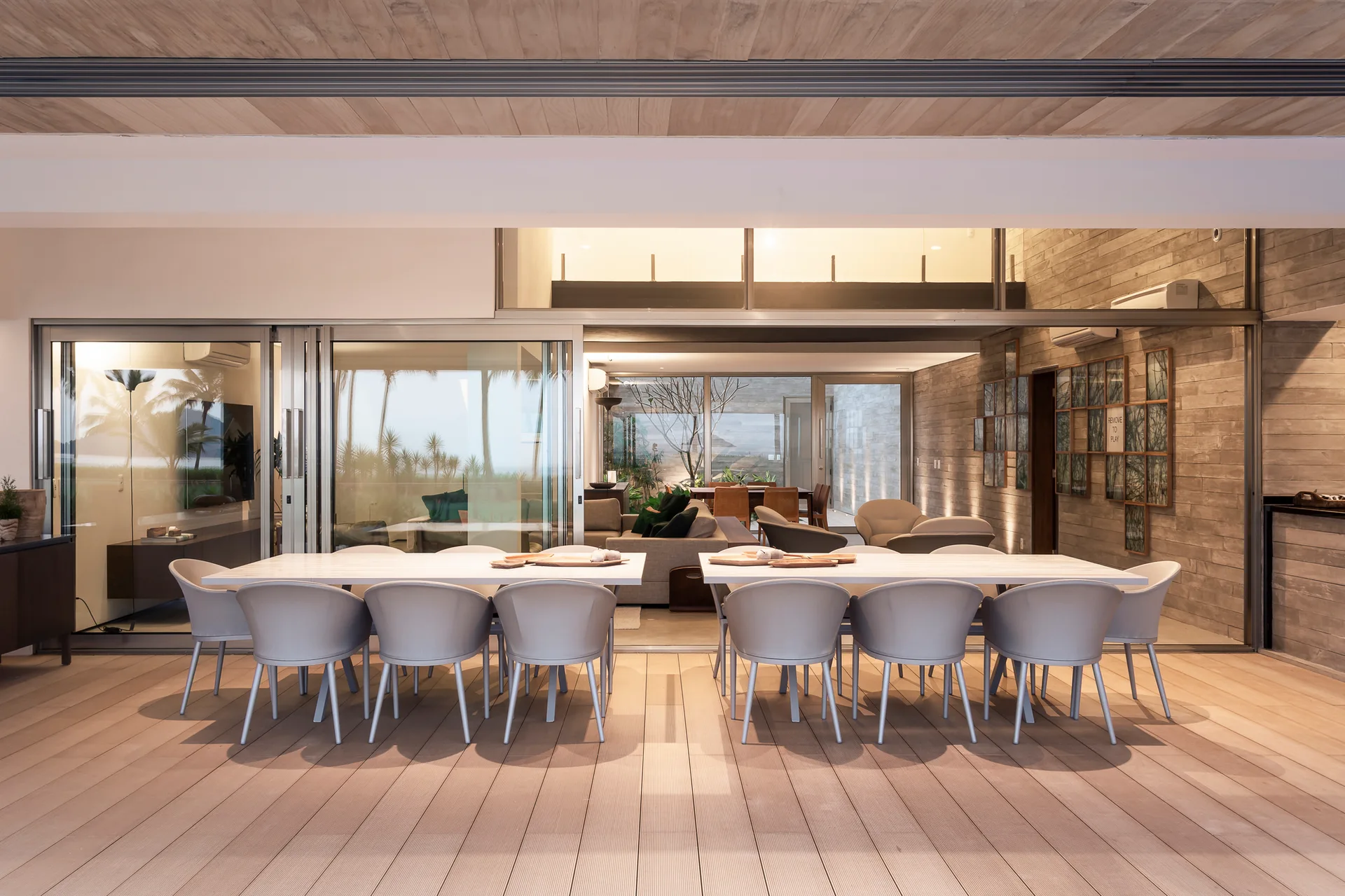 A covered outdoor patio with two long white dining tables and gray chairs, opening to an indoor living area through large sliding glass doors.