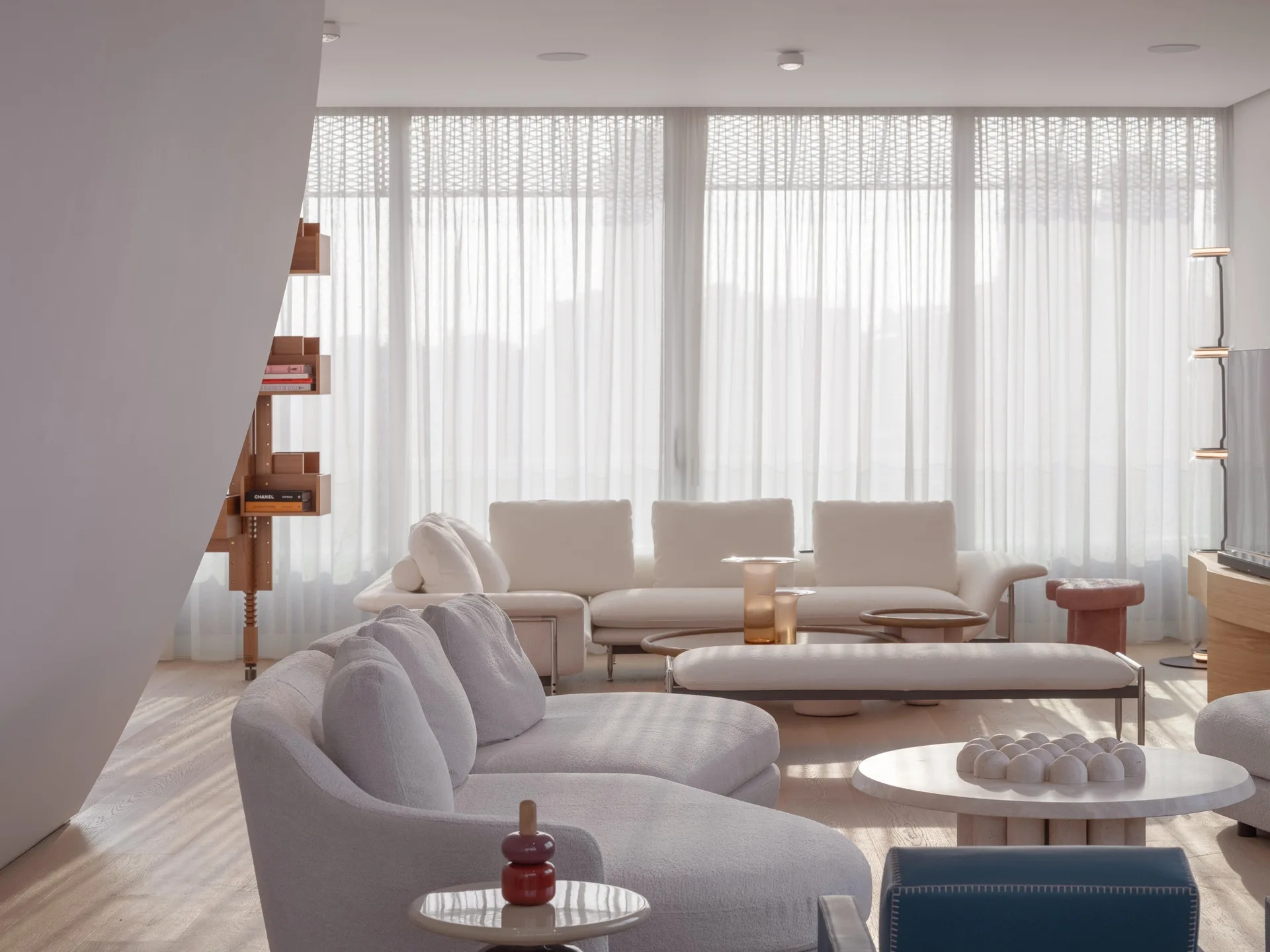 A light-filled living room features cream-colored curved sofas, floor-to-ceiling sheer curtains, a low wooden shelf, and sculptural stone coffee tables.