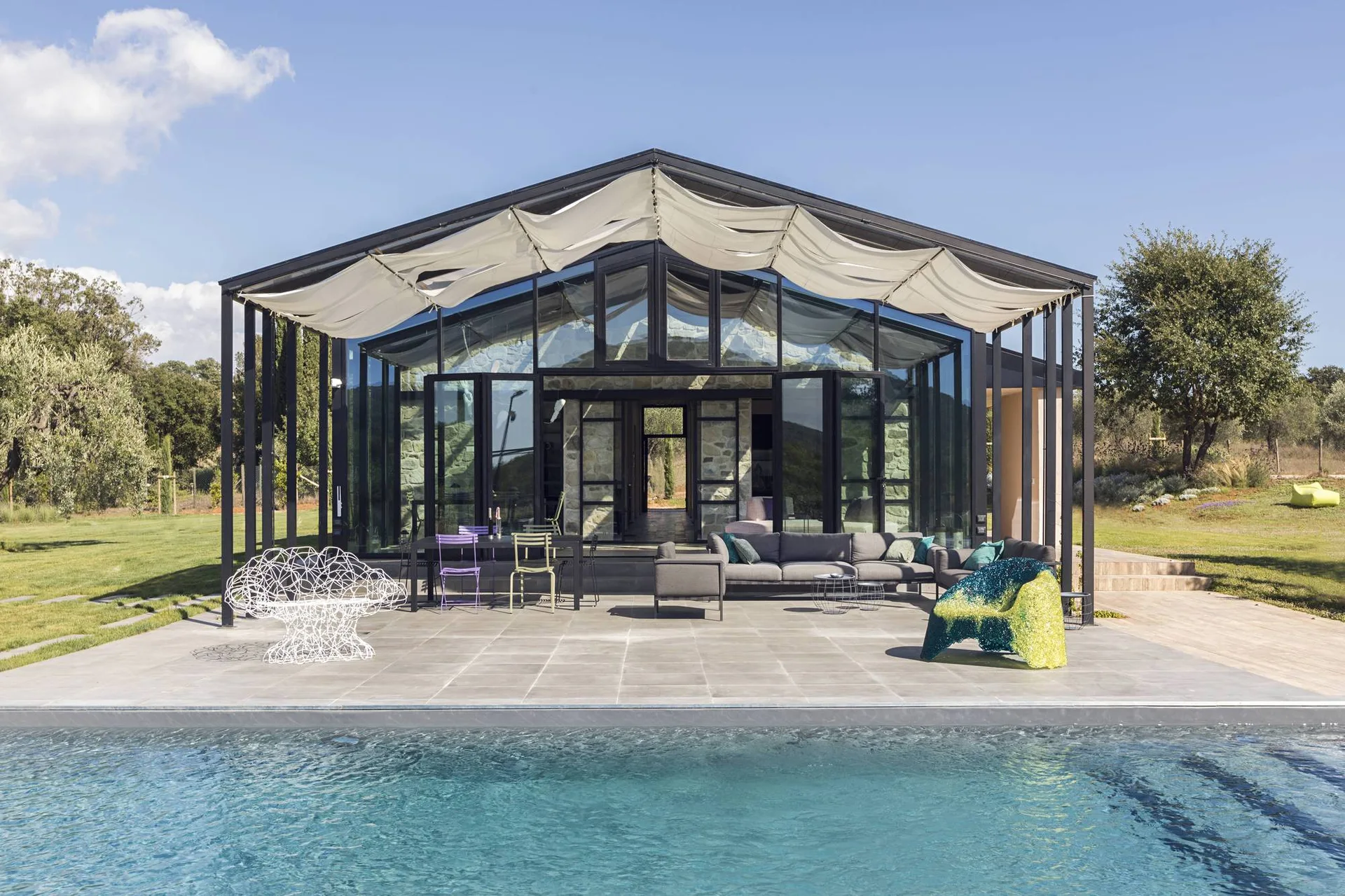 A modern glass-walled pool pavilion features a gray tiled patio with a sectional sofa, colorful accent chairs, a draped white awning, and a swimming pool.