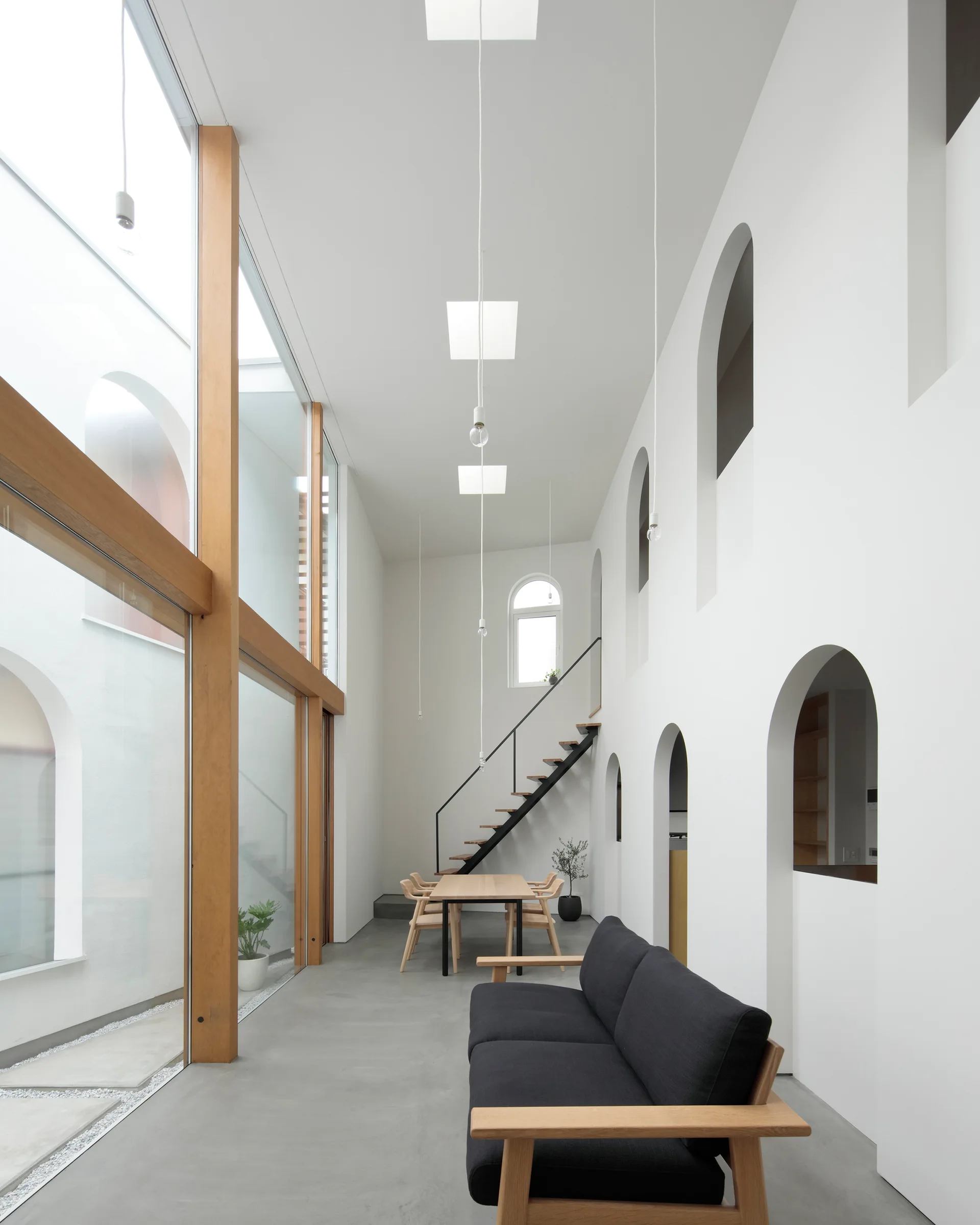 A double-height minimalist room features a black sofa, a light wood dining set, large wood-framed glass walls, and a white wall with arched openings.