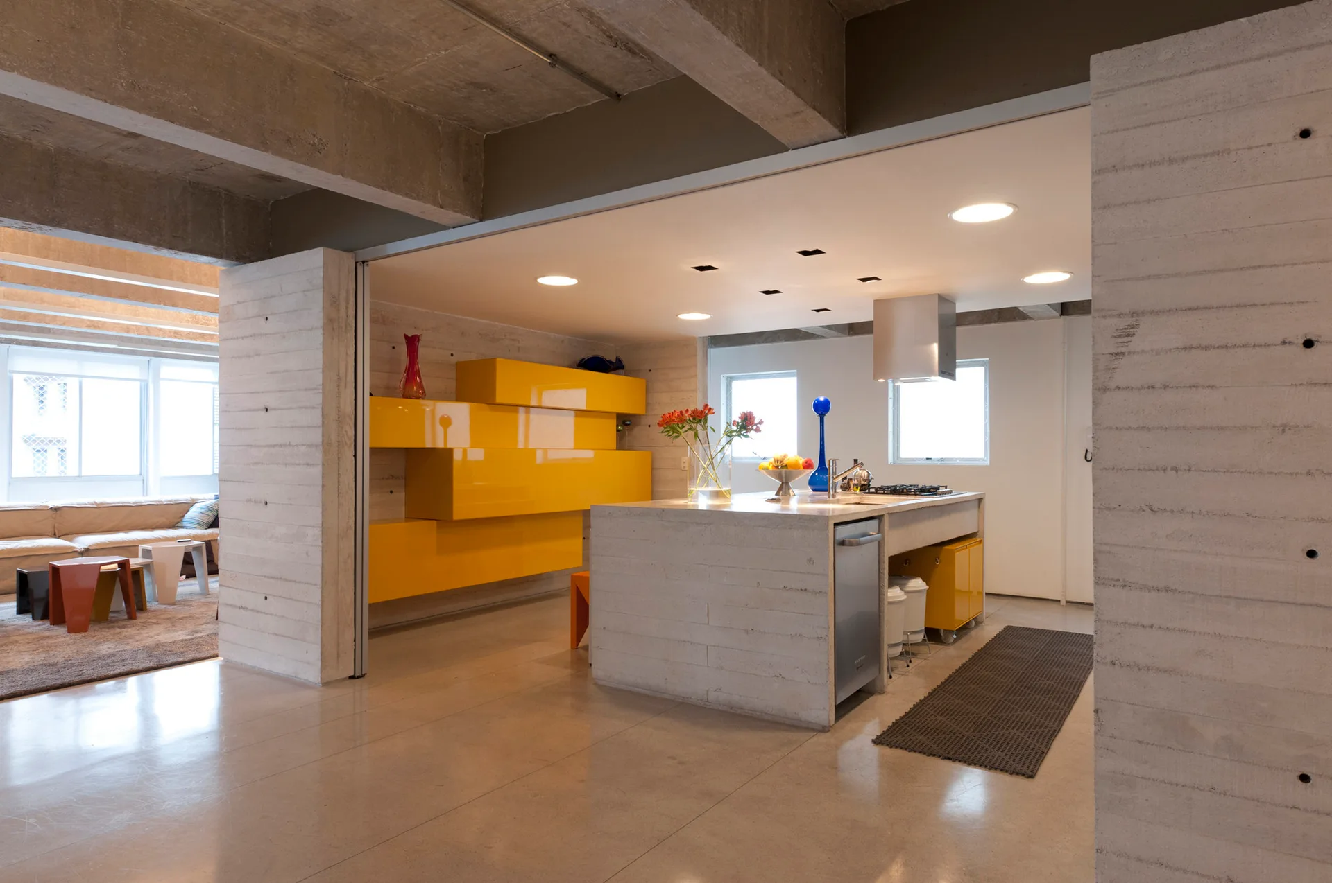 A modern kitchen features a raw concrete island, glossy yellow cabinetry, and exposed concrete walls and ceiling.