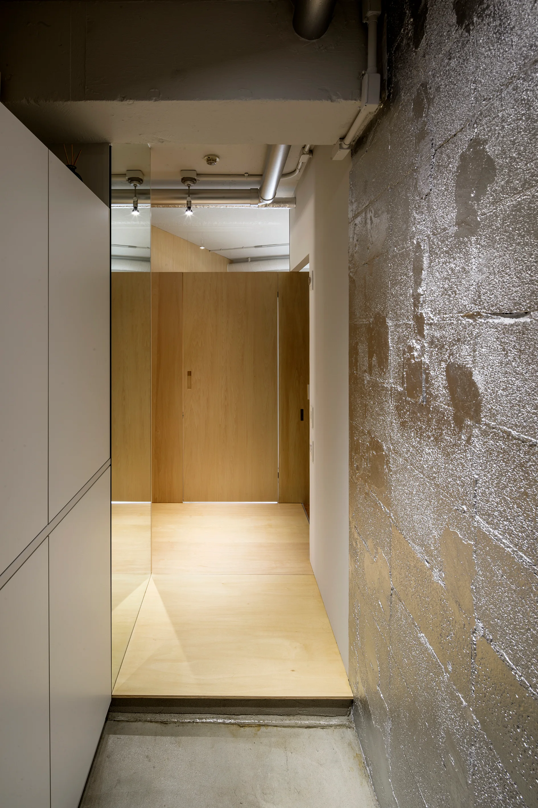 A narrow modern hallway featuring a silver-painted concrete block wall, light wood paneling, a floor-to-ceiling mirror, and white cabinets.