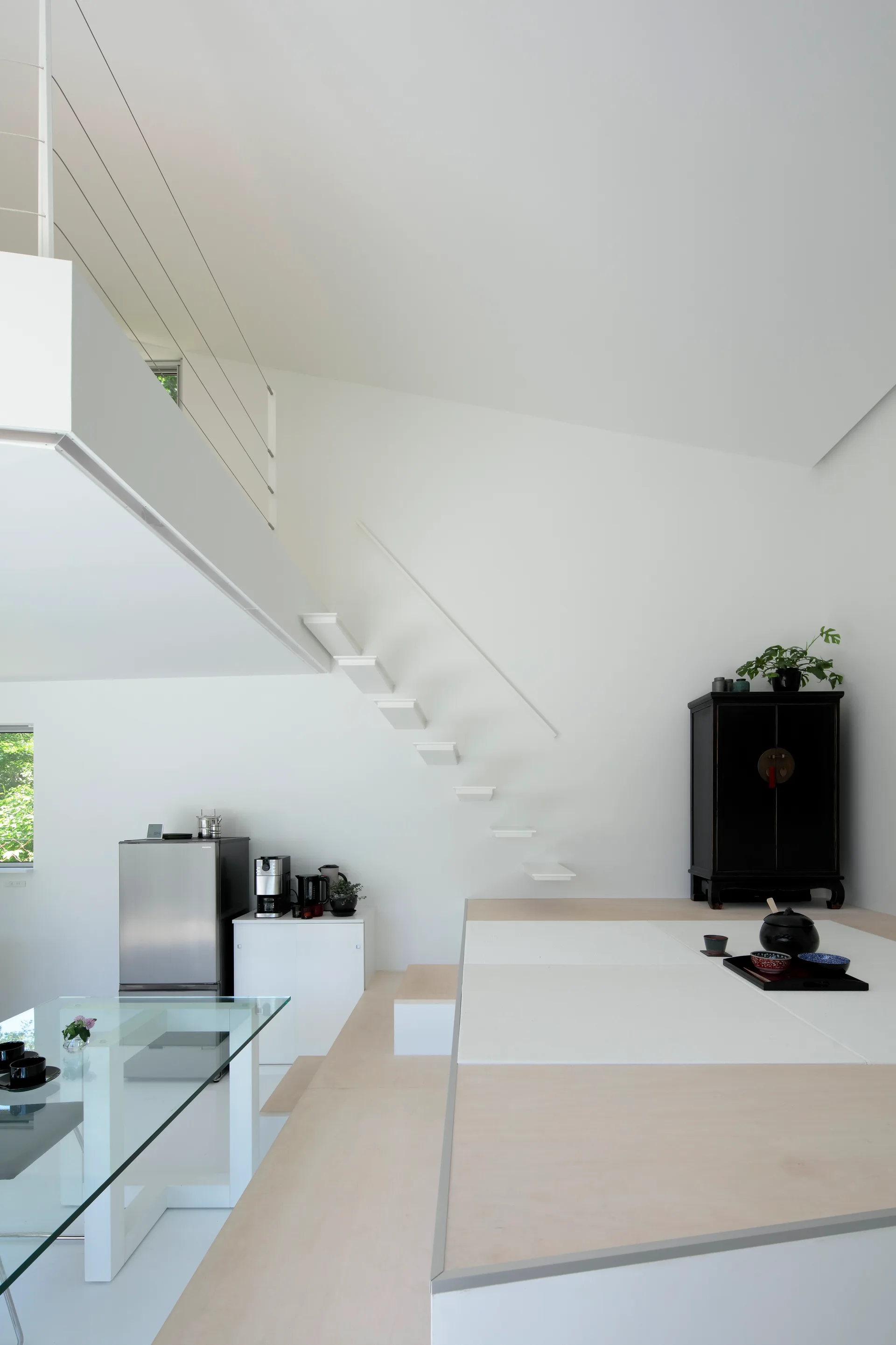 A modern split-level room features white floating stairs, a raised wood platform with a tea set, and a glass dining table.