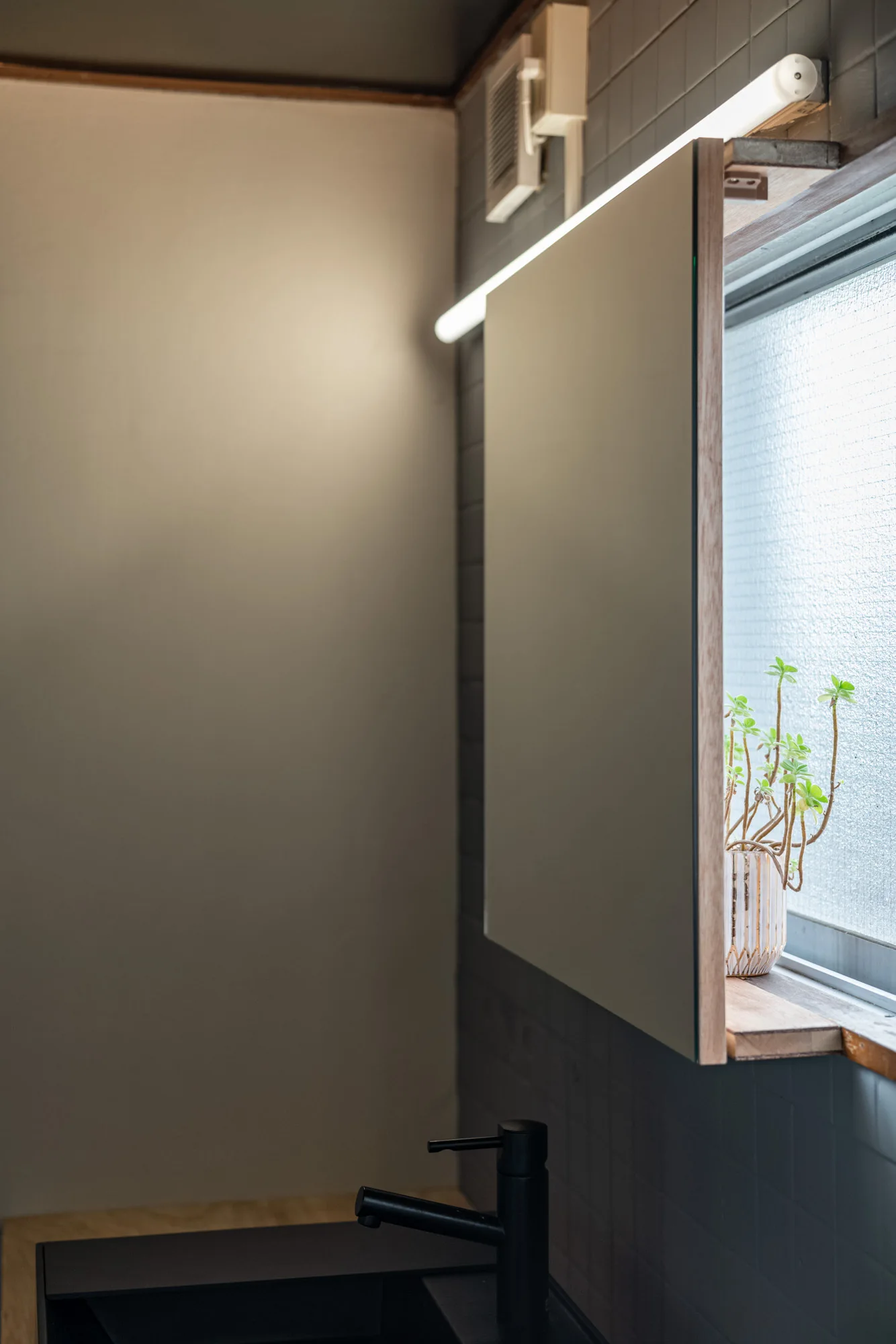 A bathroom vanity features a mirror mounted in front of a frosted glass window next to a dark grey tiled wall with a matte black faucet.