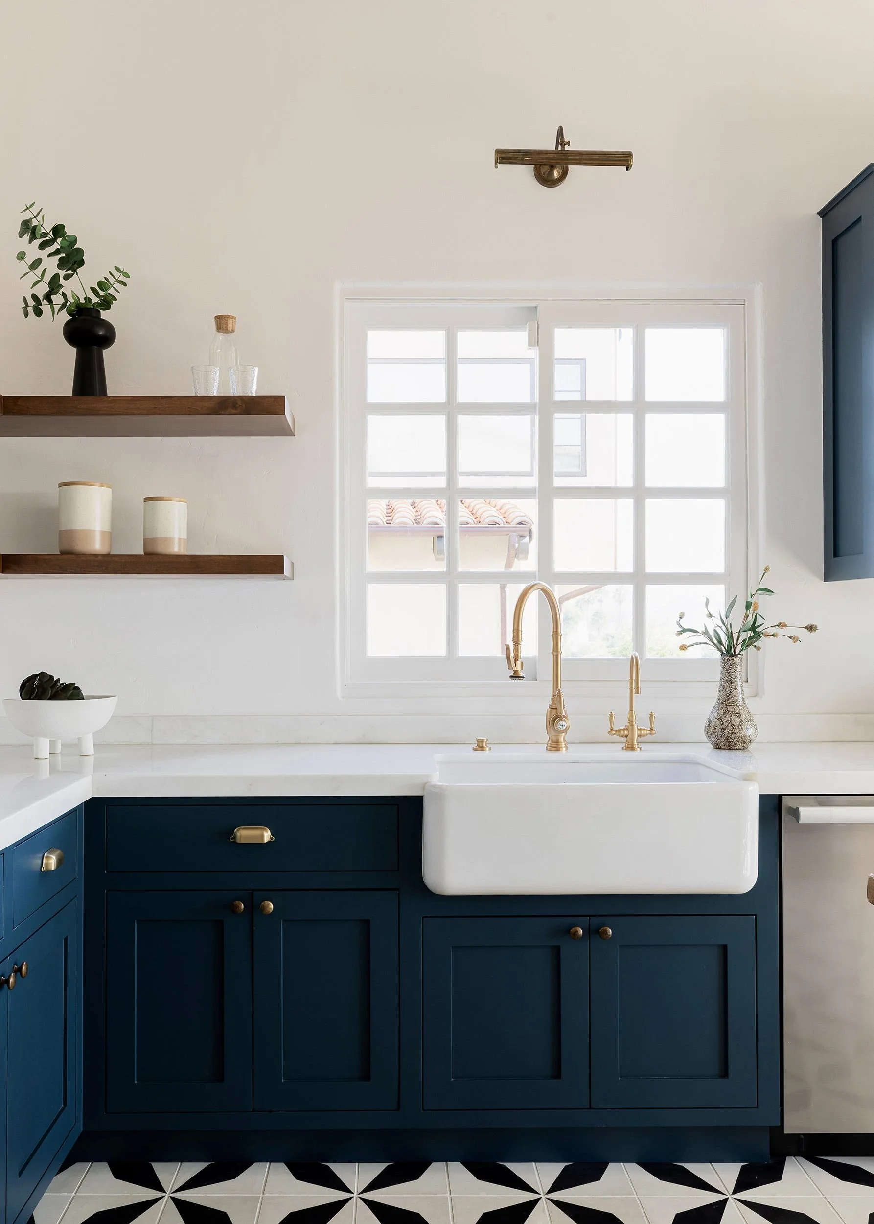 A bright kitchen features navy blue base cabinets beneath a white apron-front sink, brass faucets, and two wood floating shelves beside a grid window.