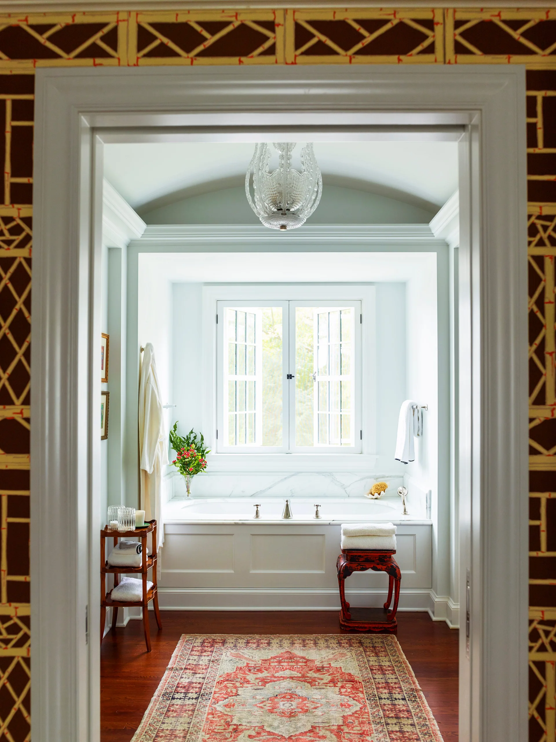 A view through a doorway of a bathroom featuring a white marble bathtub under a window, pale blue walls, and a patterned rug.