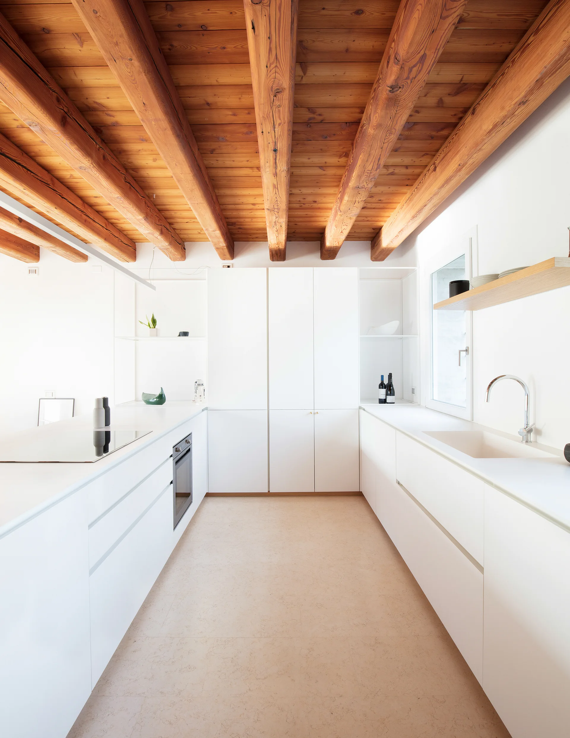 A minimalist white kitchen features flat-front cabinets, a built-in oven, and a ceiling with heavy, exposed natural wood beams.