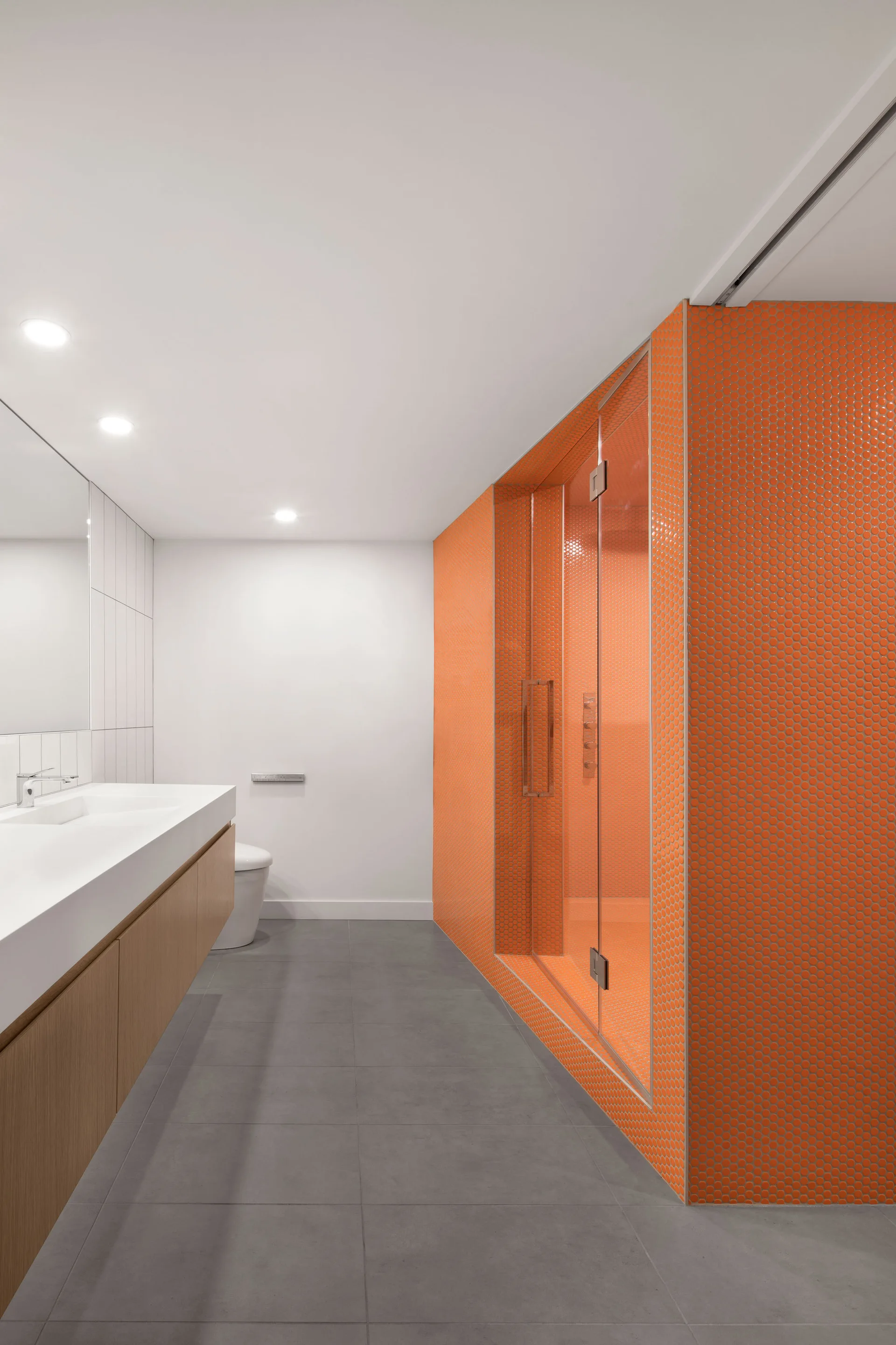 A modern bathroom features a floating wood vanity, gray floor tiles, and a glass shower lined entirely in bright orange penny tiles.