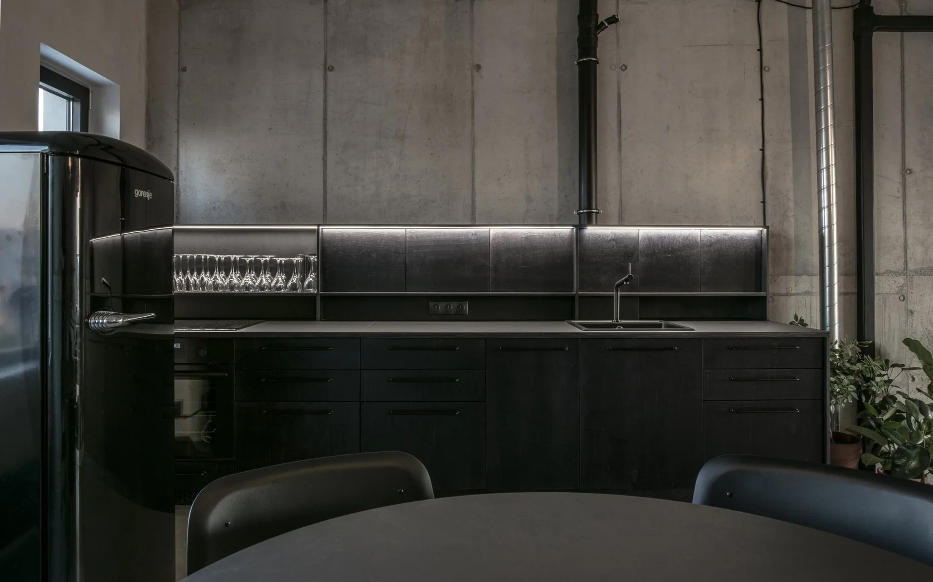 A minimalist kitchen features matte black cabinets, an integrated sink, a retro refrigerator, and an LED-backlit shelf against a raw concrete wall.