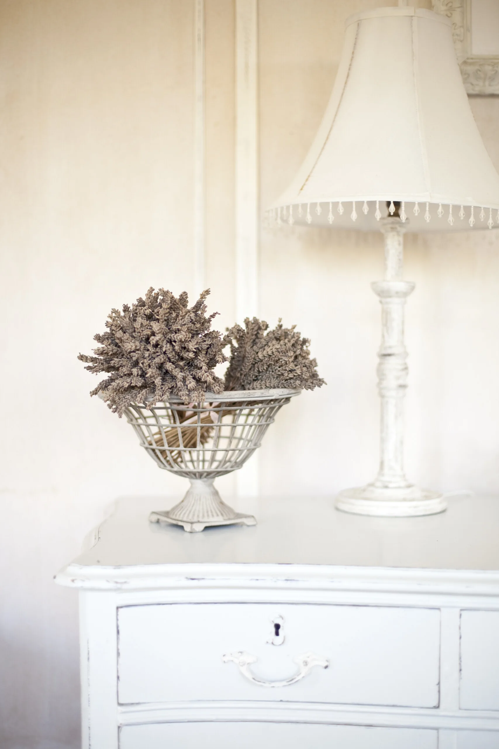 A close-up of a distressed white dresser featuring a decorative metal wire basket filled with dried lavender and a matching white table lamp.
