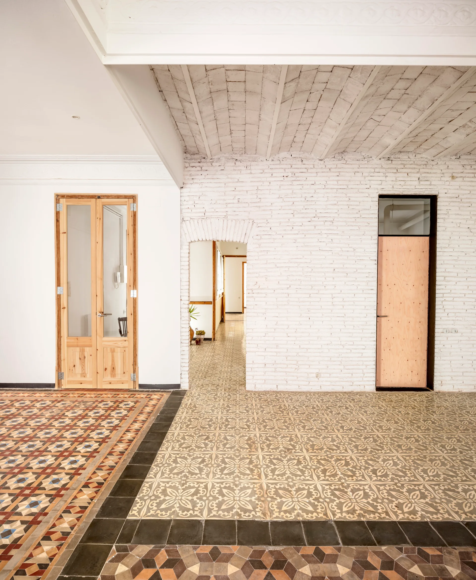An interior space features geometric patterned floor tiles, white painted brick walls, a vaulted brick ceiling, and natural wood doors.