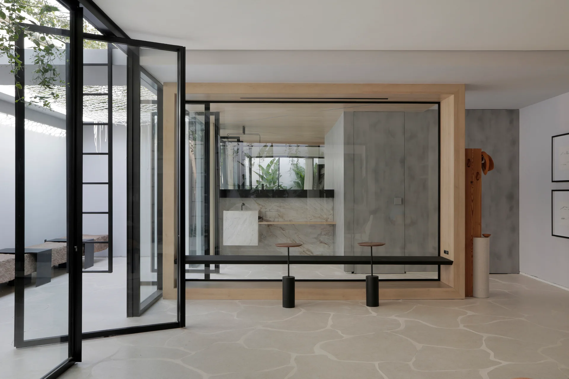A minimalist interior with a light wood framed glass bathroom partition enclosing a marble vanity next to large black-framed glass doors.