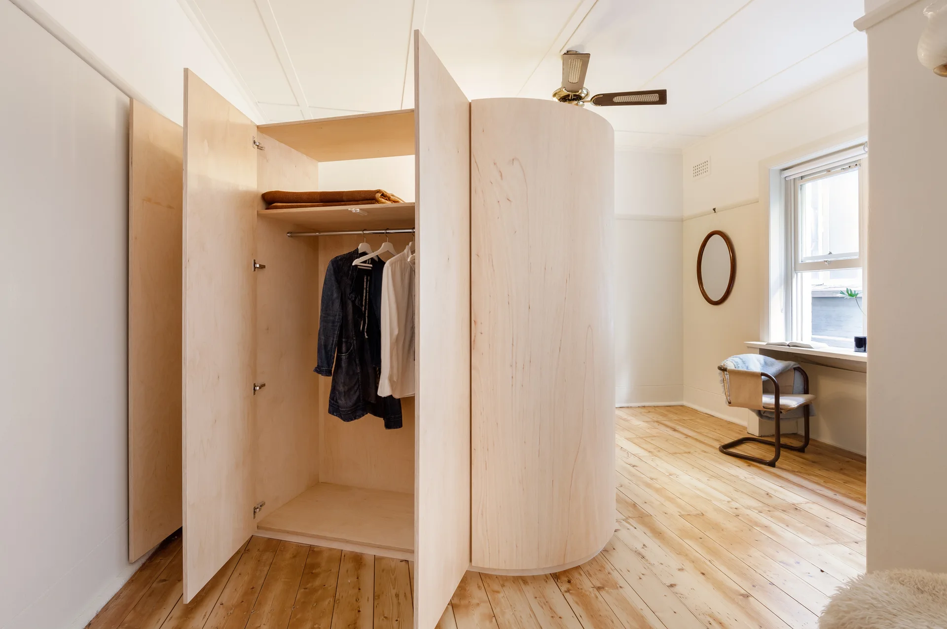 A bedroom features a central custom plywood wardrobe with open doors, wood plank flooring, a window desk, and an oval wall mirror.