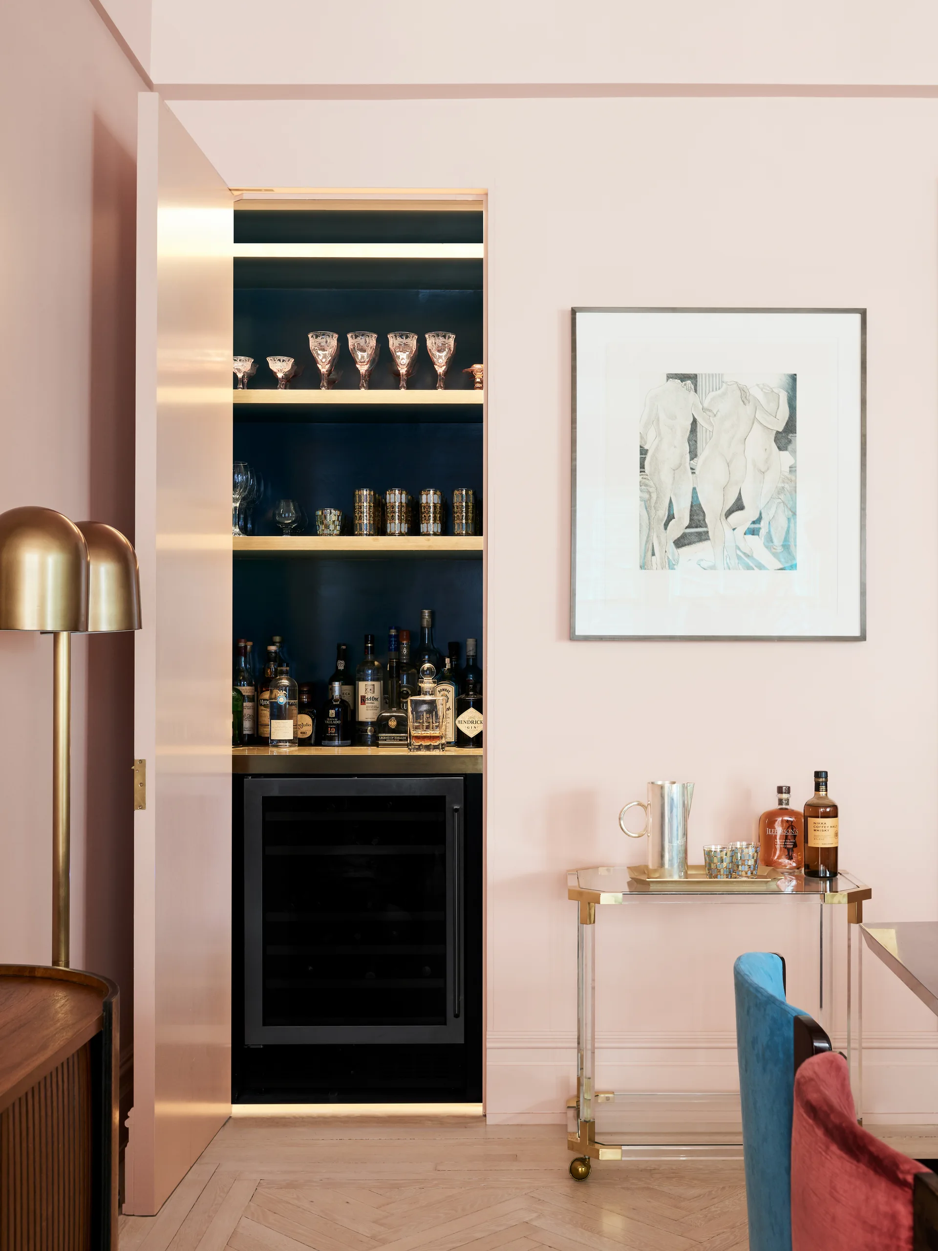 A built-in home bar with navy blue shelving and a wine cooler is revealed behind an open blush pink door in a dining area.