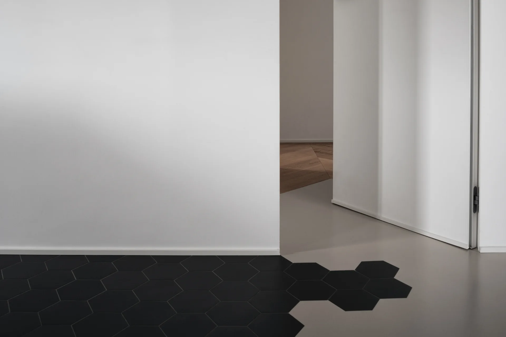 A minimalist floor transition shows matte black hexagonal tiles blending into a smooth gray surface next to a white wall and an open doorway.