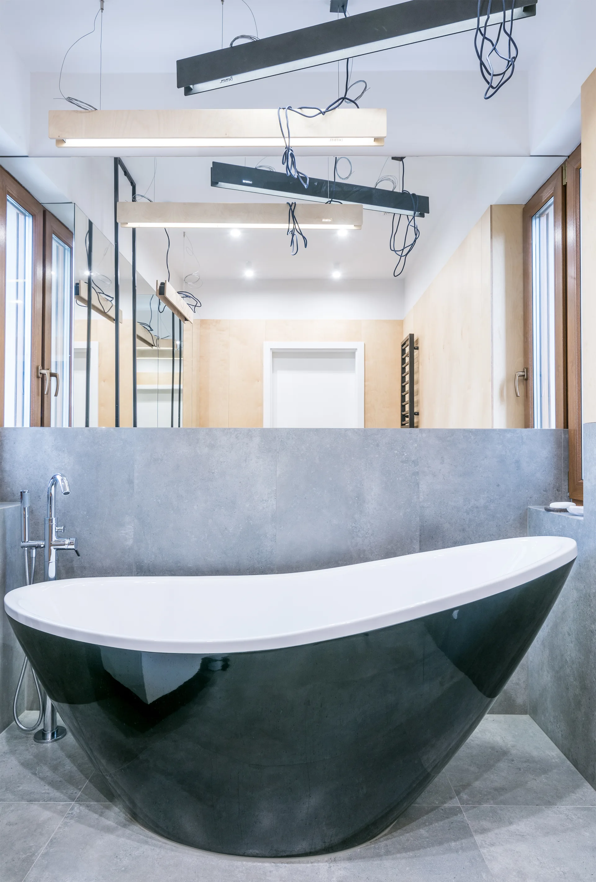 A modern bathroom features a glossy black-and-white freestanding bathtub set against large grey tiles, topped by a large wall mirror and linear pendant lights.