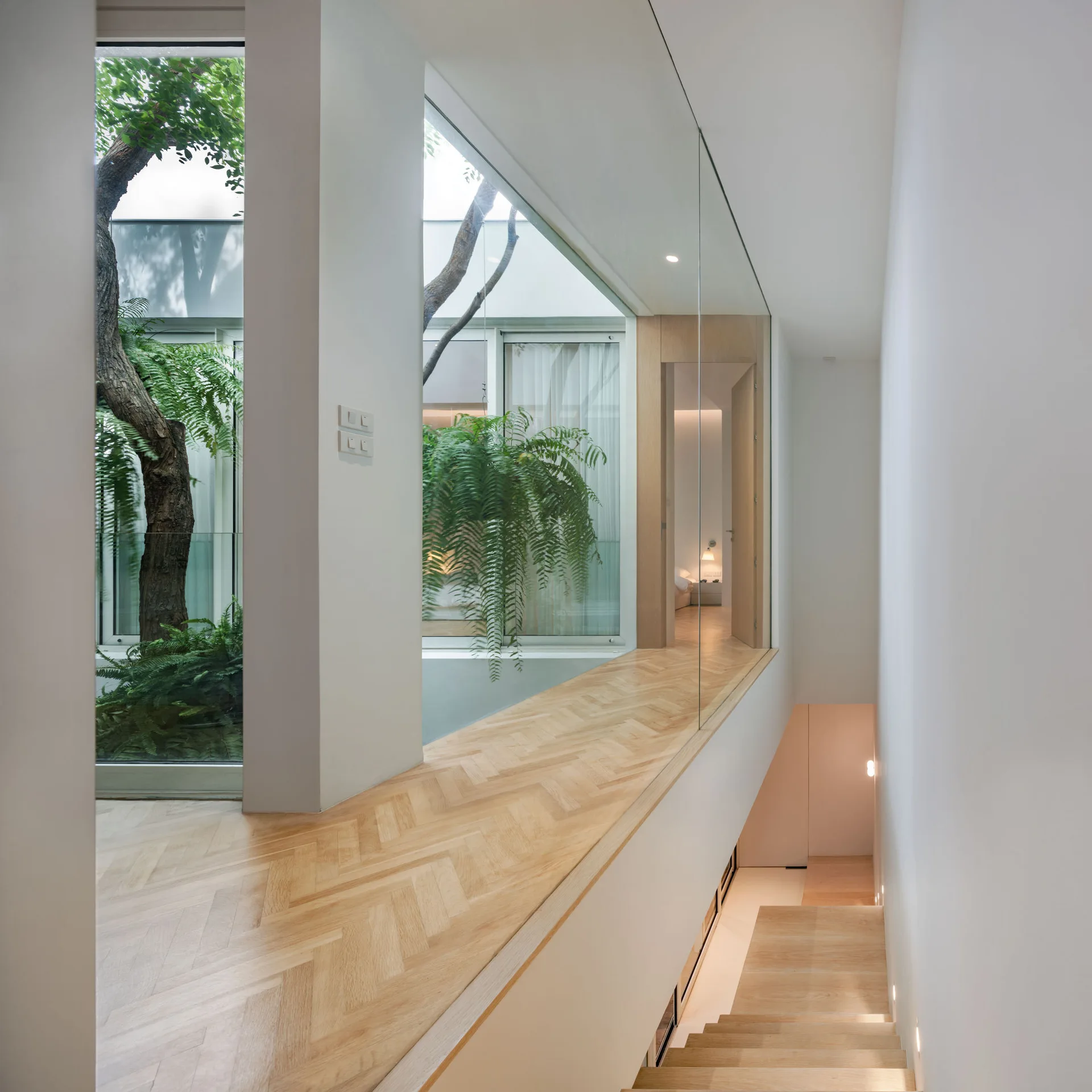 A minimalist hallway featuring light wood herringbone flooring and a clear glass wall overlooking an indoor courtyard with green plants.