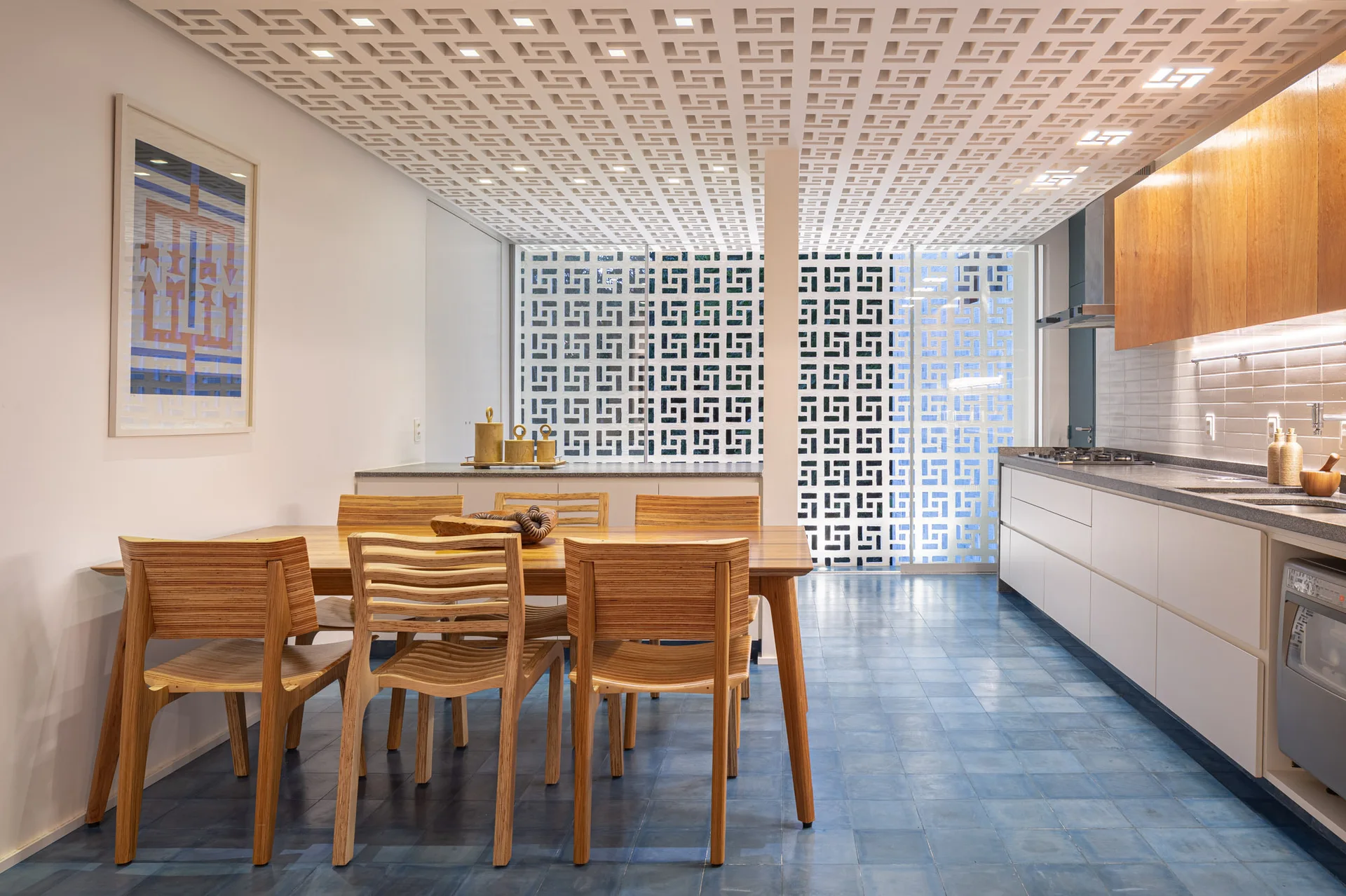 A modern kitchen and dining area with a wood table, slatted chairs, blue floor tiles, and a white geometric ceiling screen.