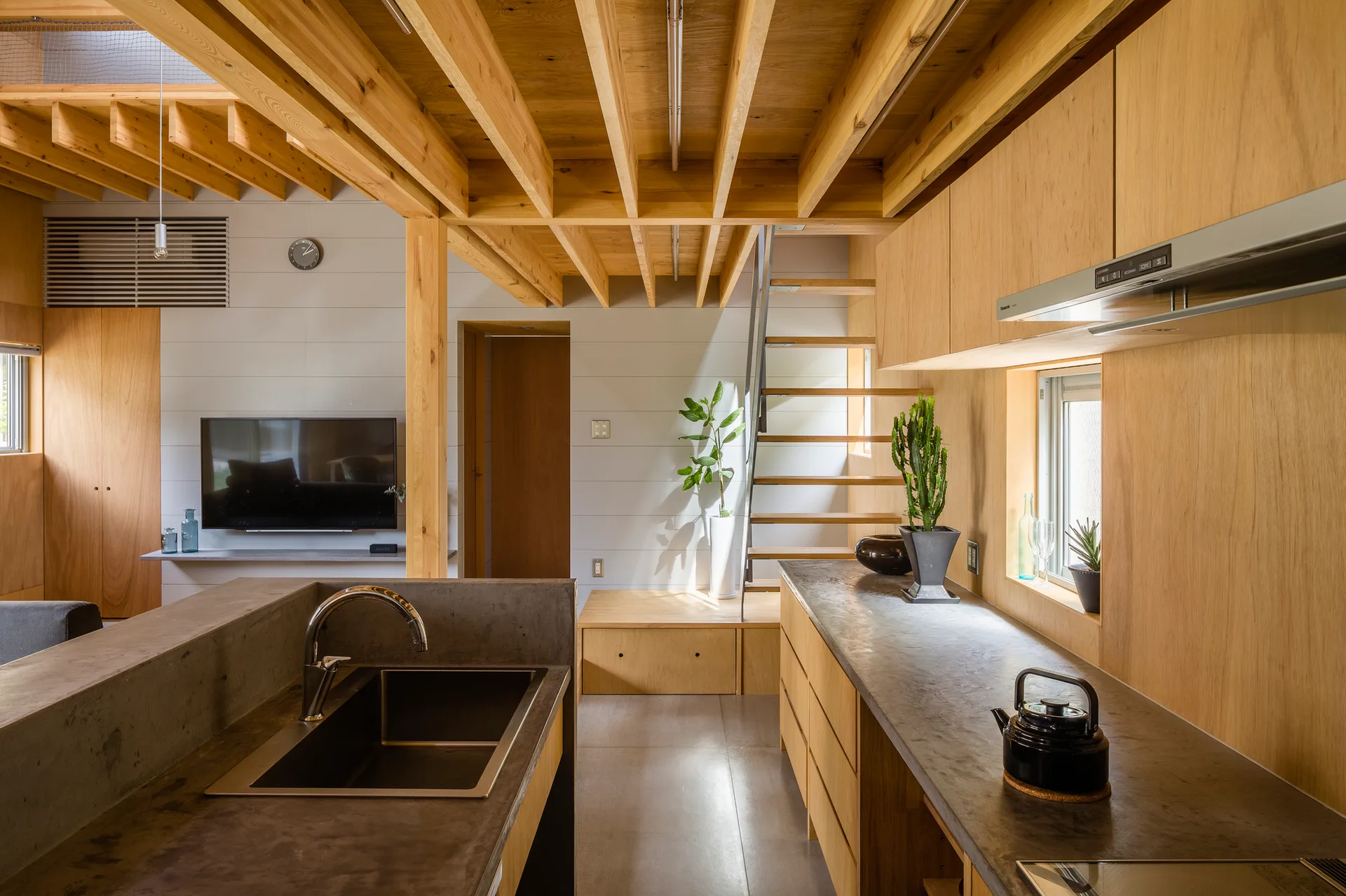 A modern open-plan kitchen featuring exposed wooden ceiling beams, concrete countertops, wood veneer cabinetry, and an open riser staircase.