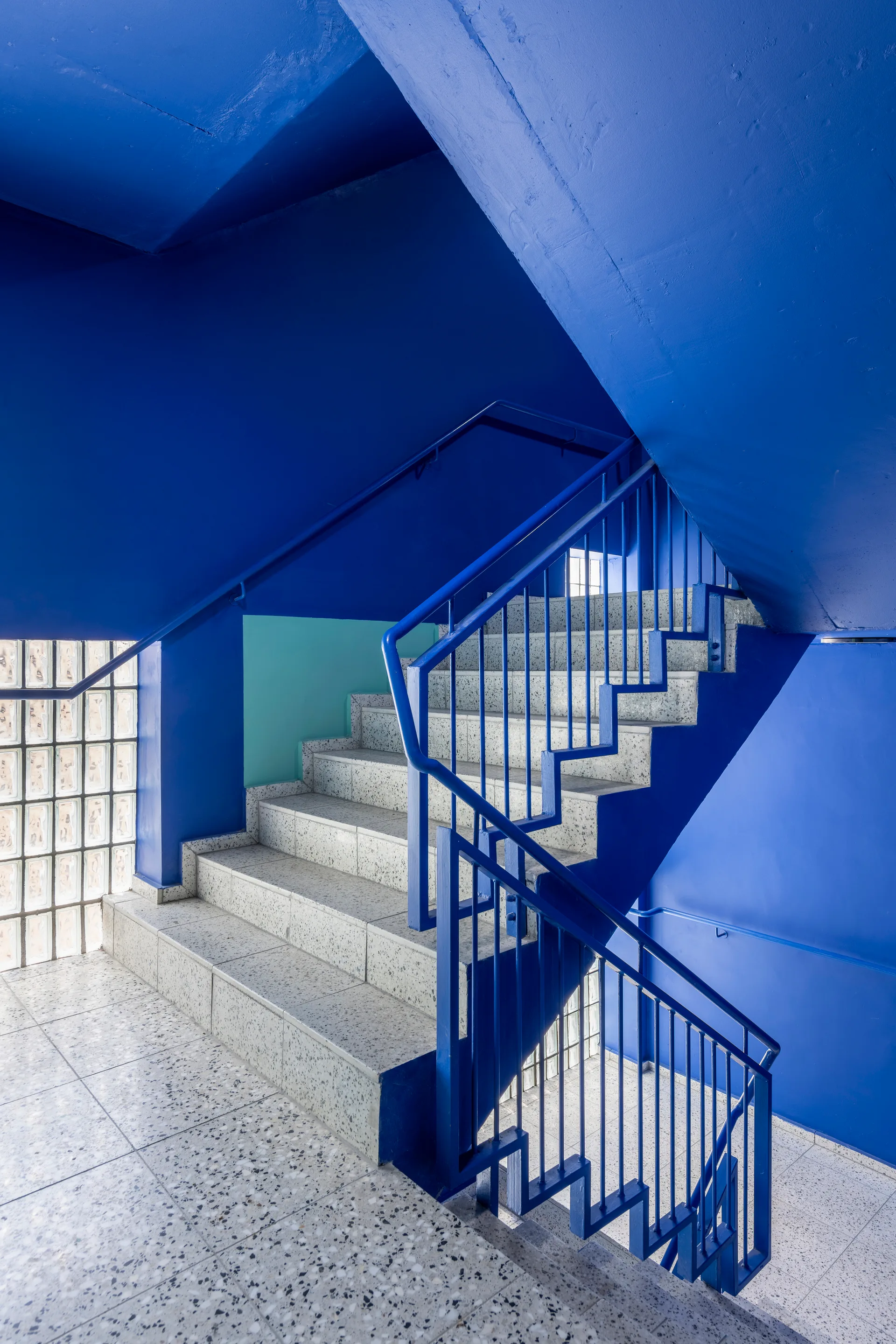 A blue stairwell features terrazzo stone steps, matching blue metal railings, and a grid-like glass block wall.