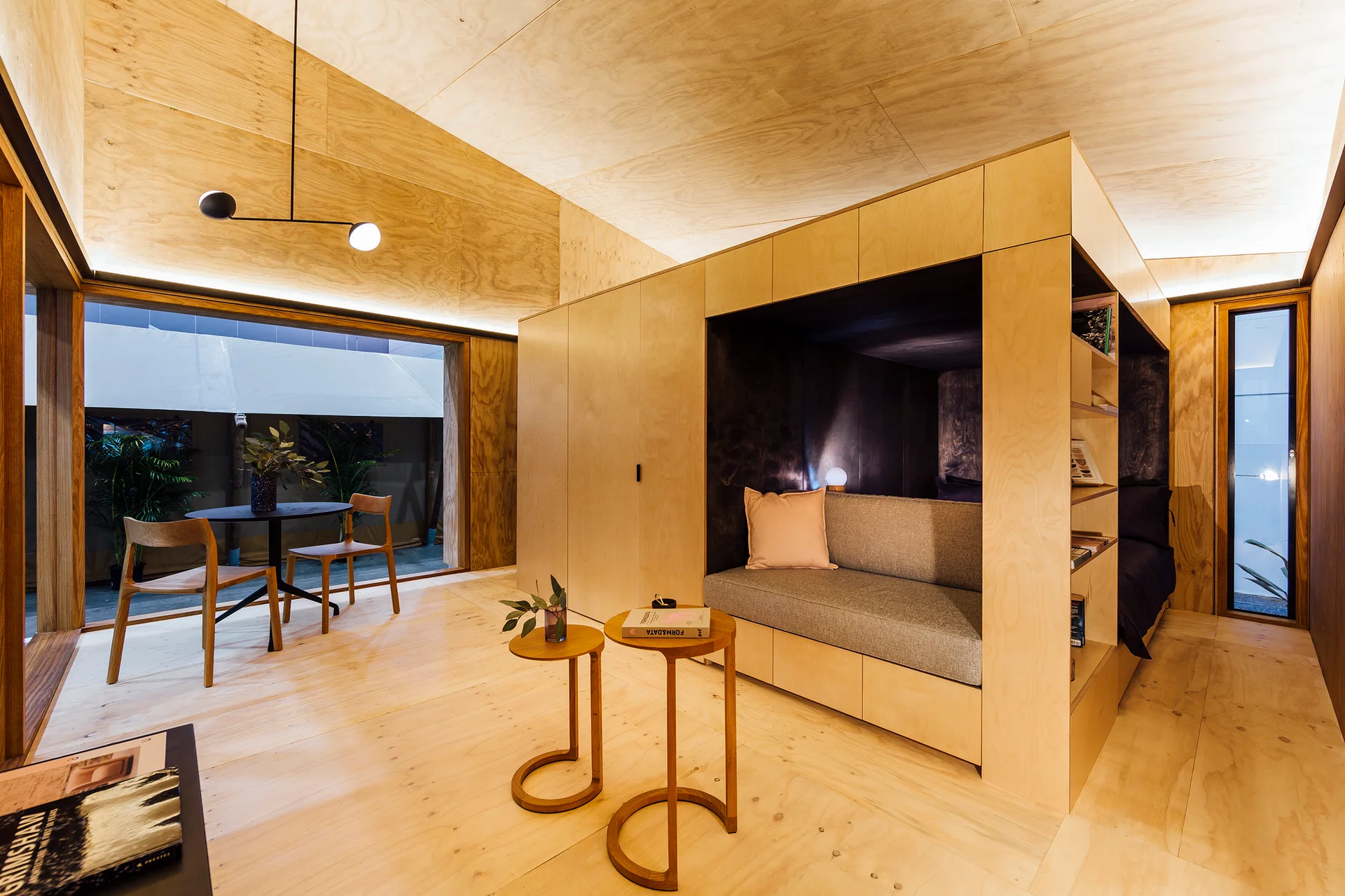 A minimalist wooden bedroom features light plywood walls and ceiling surrounding a large central built-in storage unit with an integrated sofa nook.